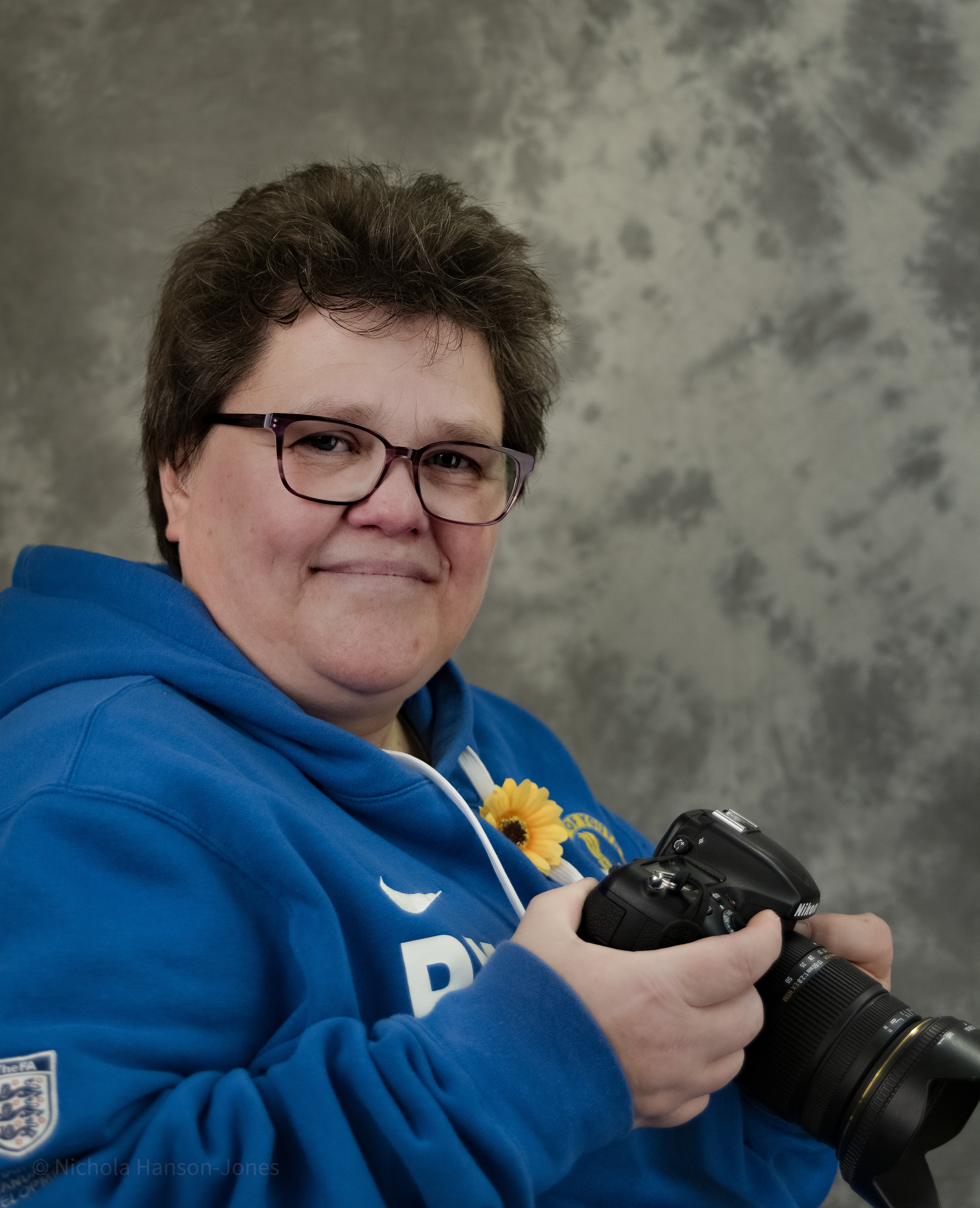 A portrait of a woman, wearing a blue football top, with a yellow sunflower pinned to her, she is holding a camera in her hands, and she is looking at the photographer and smiling.