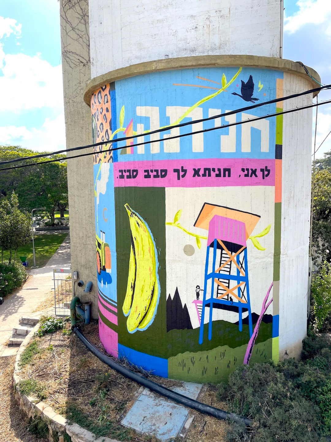 Colorful mural on a cylindrical building depicts a yellow banana, blue tower, and black bird on a blue background, with Hebrew text on a pink band.