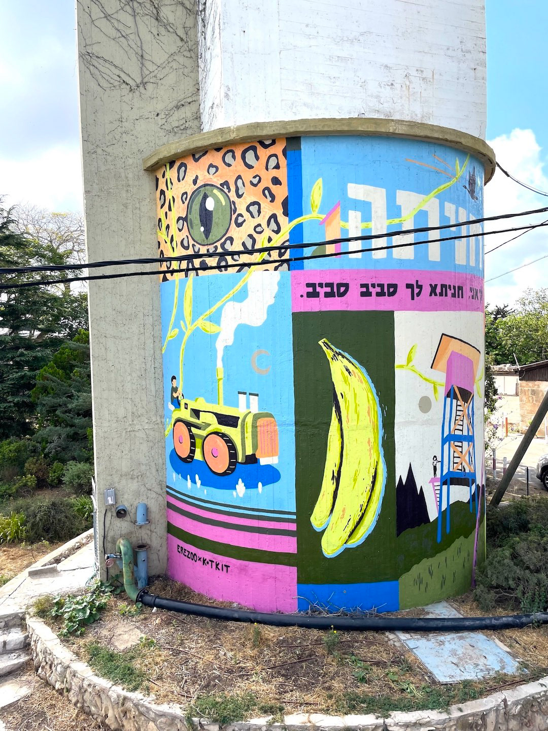 Colorful street art wraps around a cylindrical structure, featuring a leopard print eye, tractor, bananas, and a ladder. Vibrant and whimsical scene.
