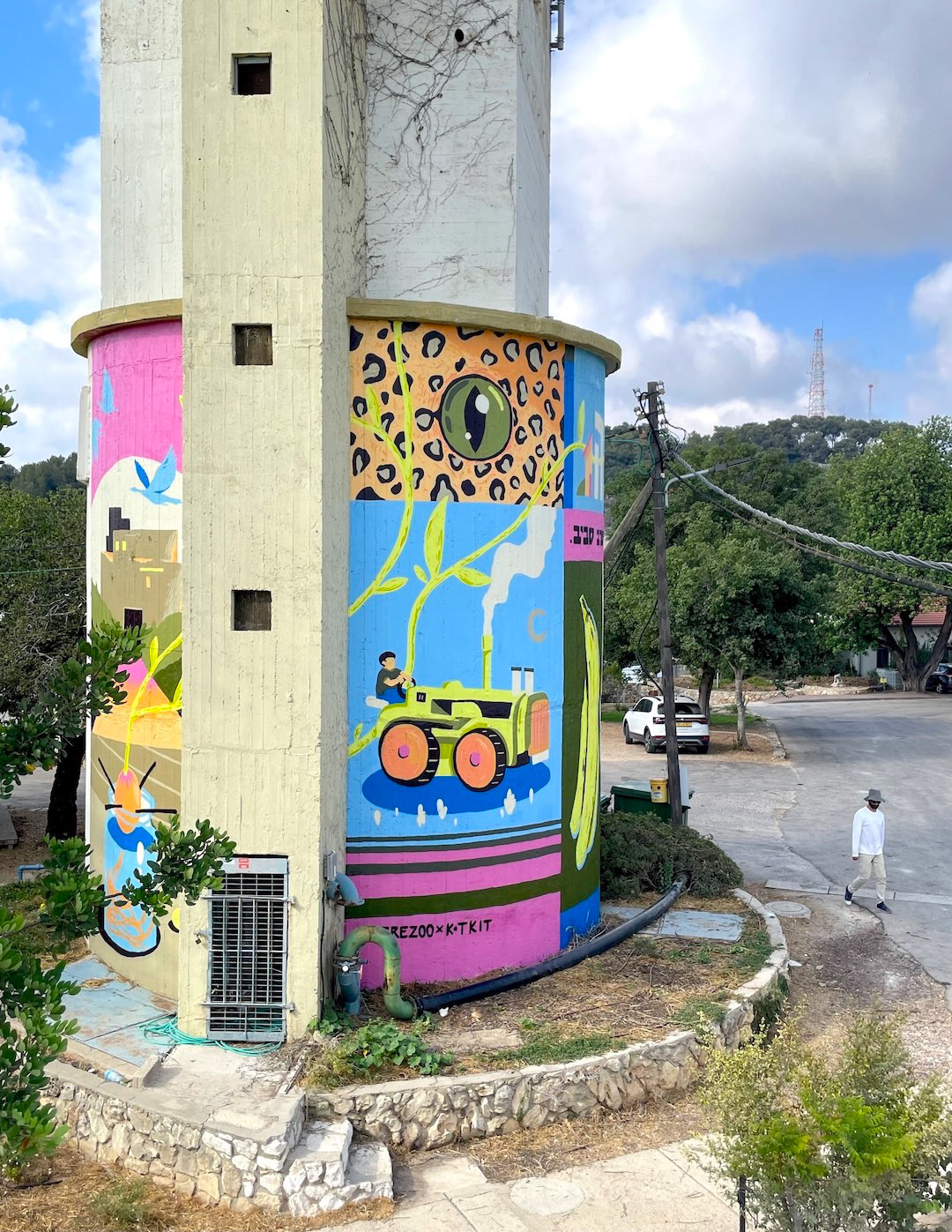 A vibrant mural wraps around a cylindrical structure, featuring bold designs like a leopard's eye, a tractor, and colorful abstract motifs. The street setting is calm, with trees and a person walking nearby, evoking a lively and artistic atmosphere.