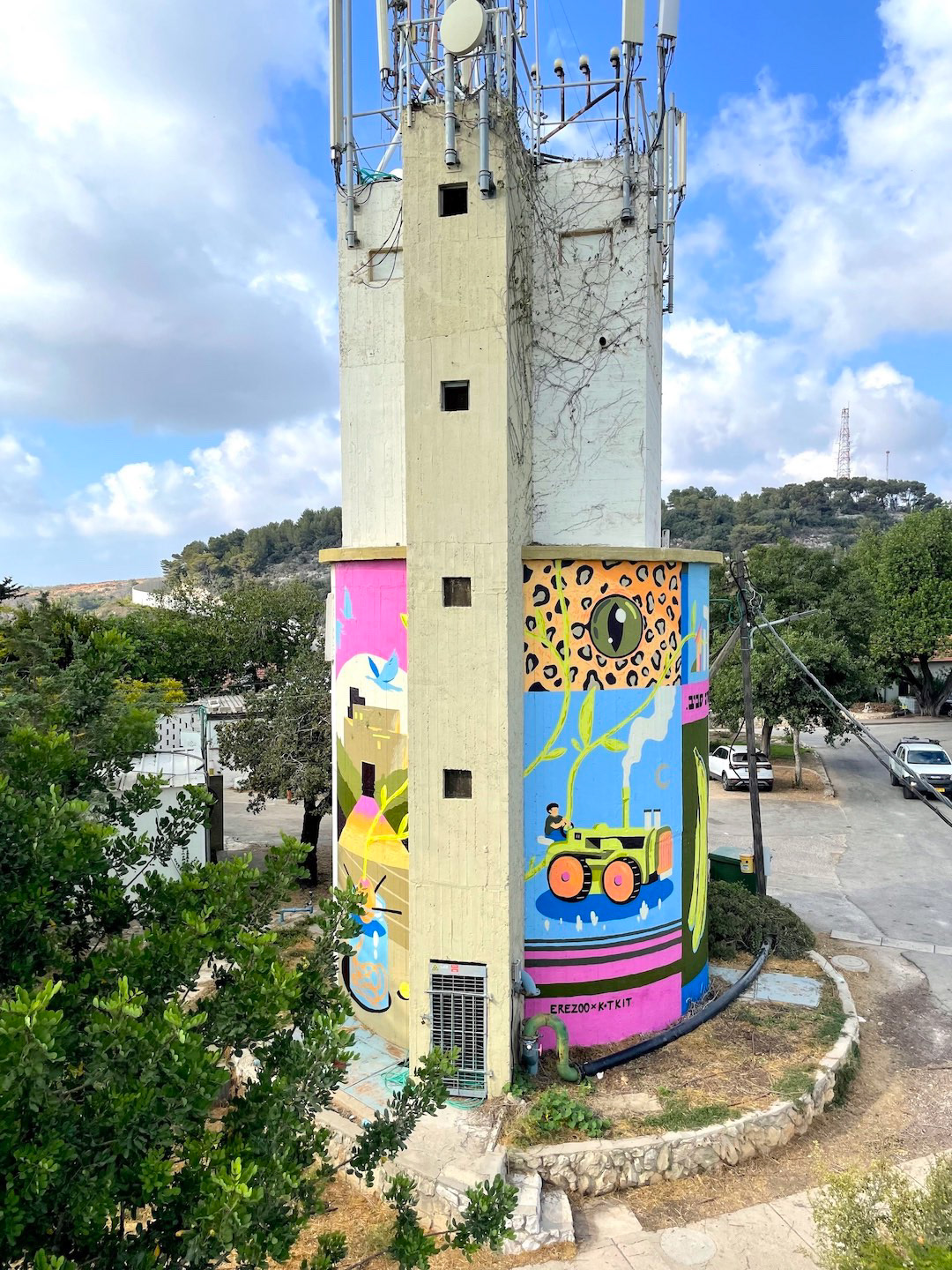A tall utility tower with antennas is painted with colorful murals, including a green tractor