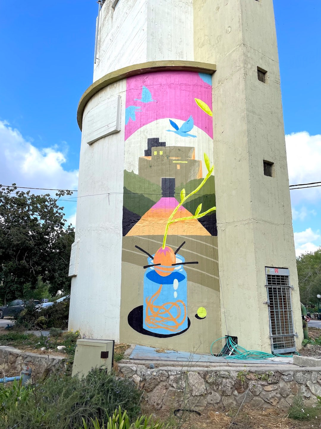 Bright mural on a cylindrical tower features abstract shapes: a jar, feathers, building, and birds. Vivid colors and a clear blue sky.