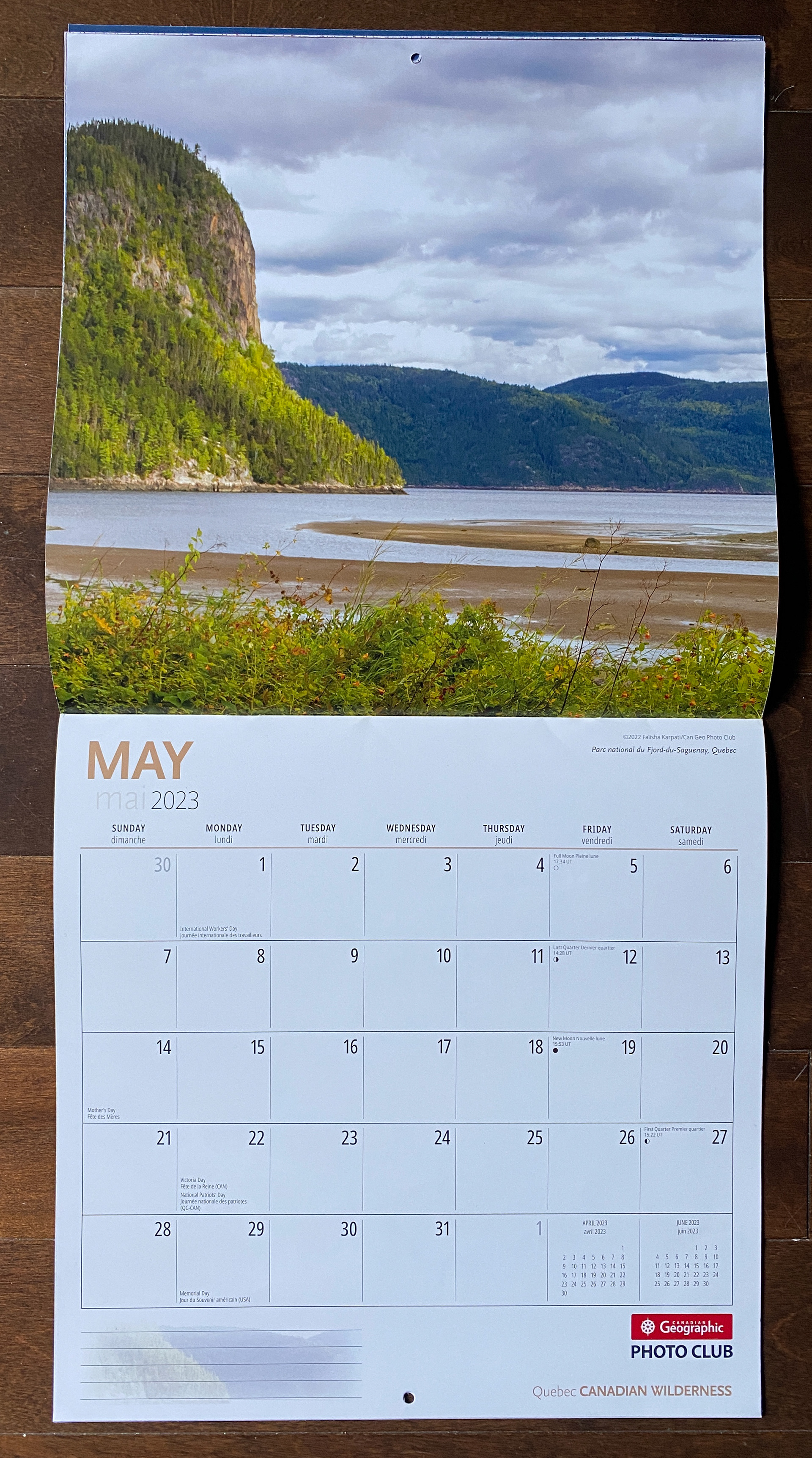 Opened calendar with May 2023 on the bottom and Falisha's photo of a large rounded cliff on the left , cloudy sky, sandy curves going into a body of water in the middle, and a line of plants at the bottom.