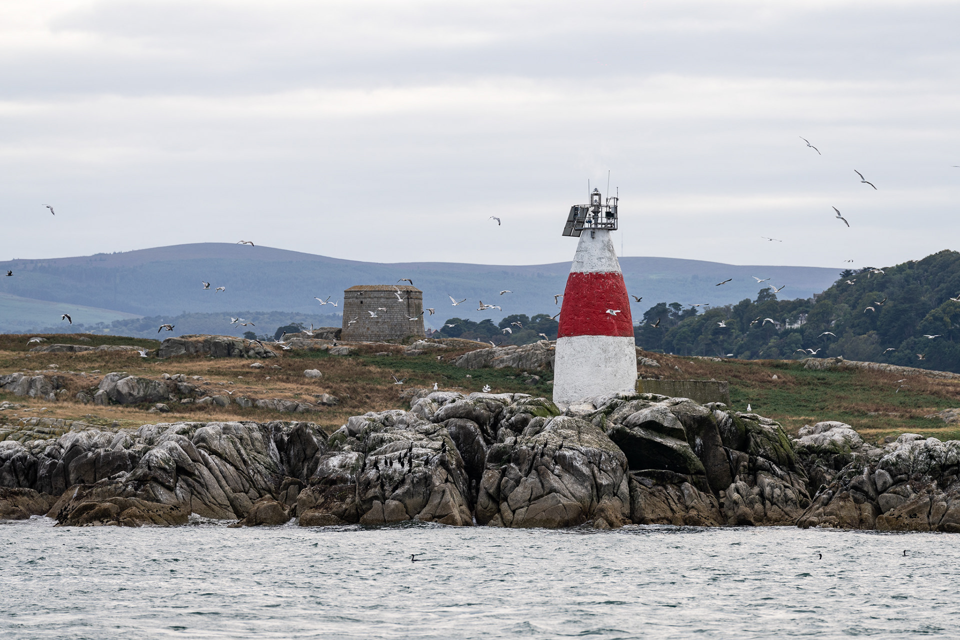 A white and red aid to navigation sits in front of and to the right of a centuries-old signal tower, where fires were once lit to warn the surrounding area of incoming dangers