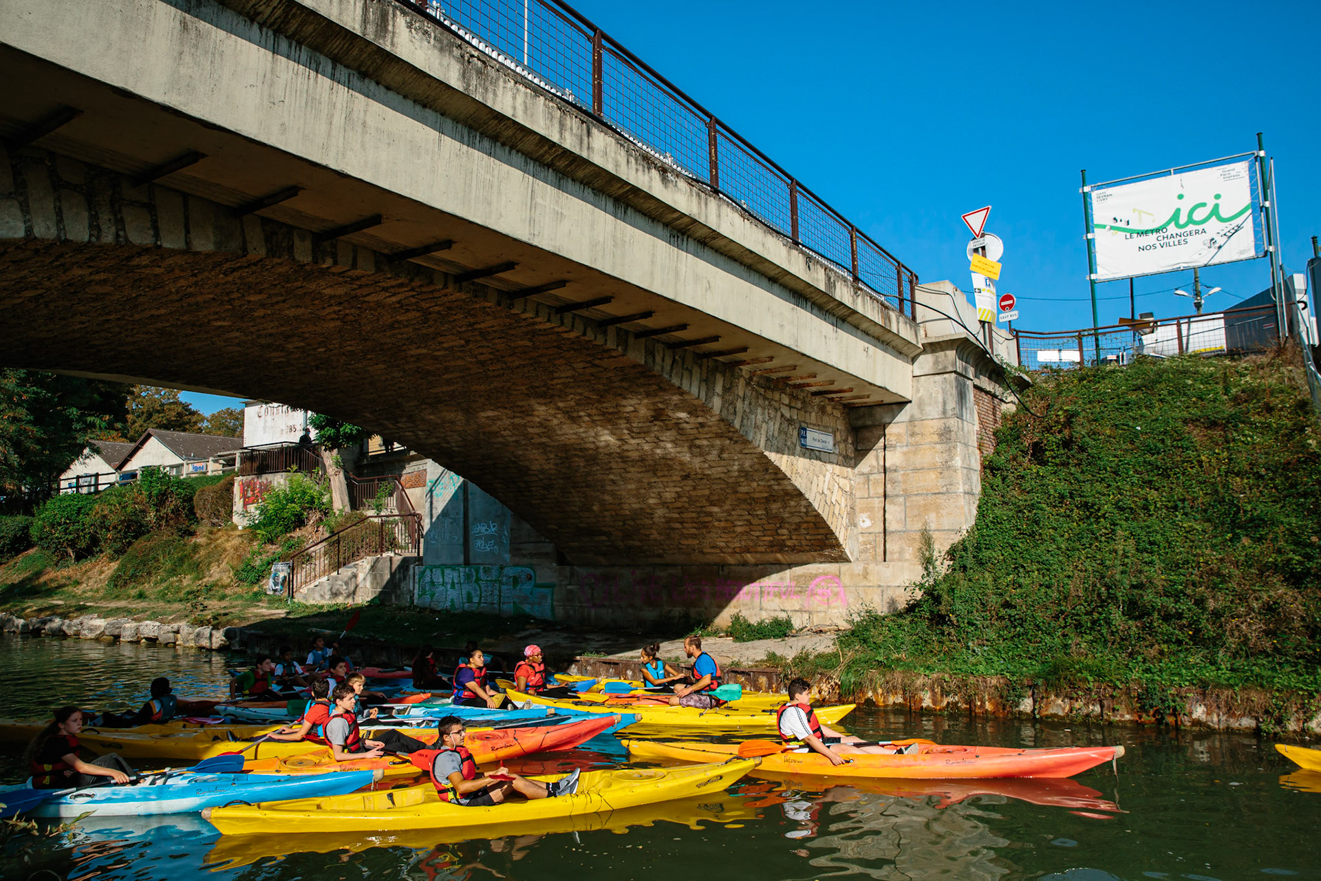Un groupe scolaire fait du kayak un bon matin ensoleillé sous le pont du centre de Sevran enjambant le canal de l’Ourcq.