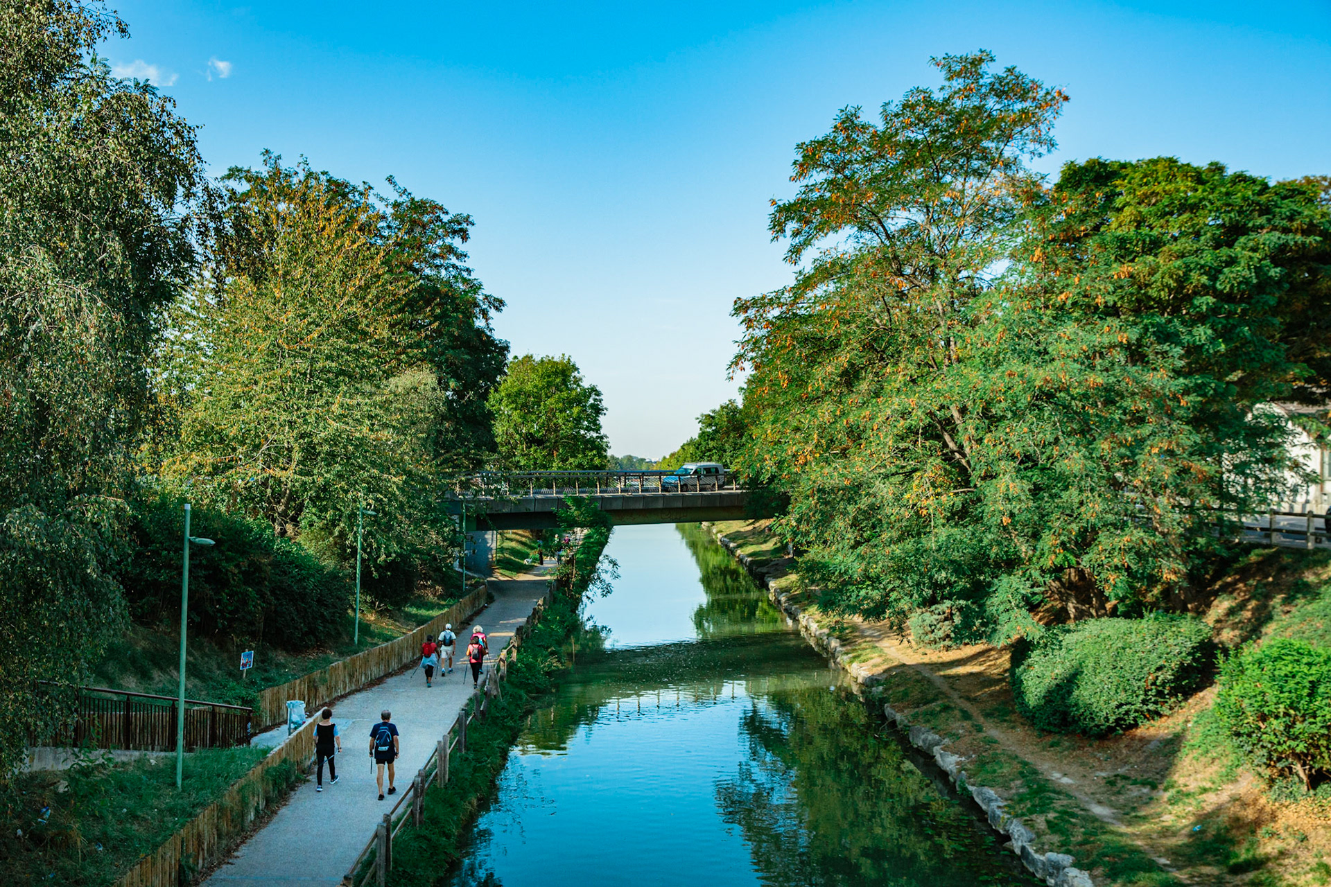 Vue du canal de l’Ourcq depuis le pont au centre de Sevran. Un groupe de personne fait de la marche nordique le long du canal.