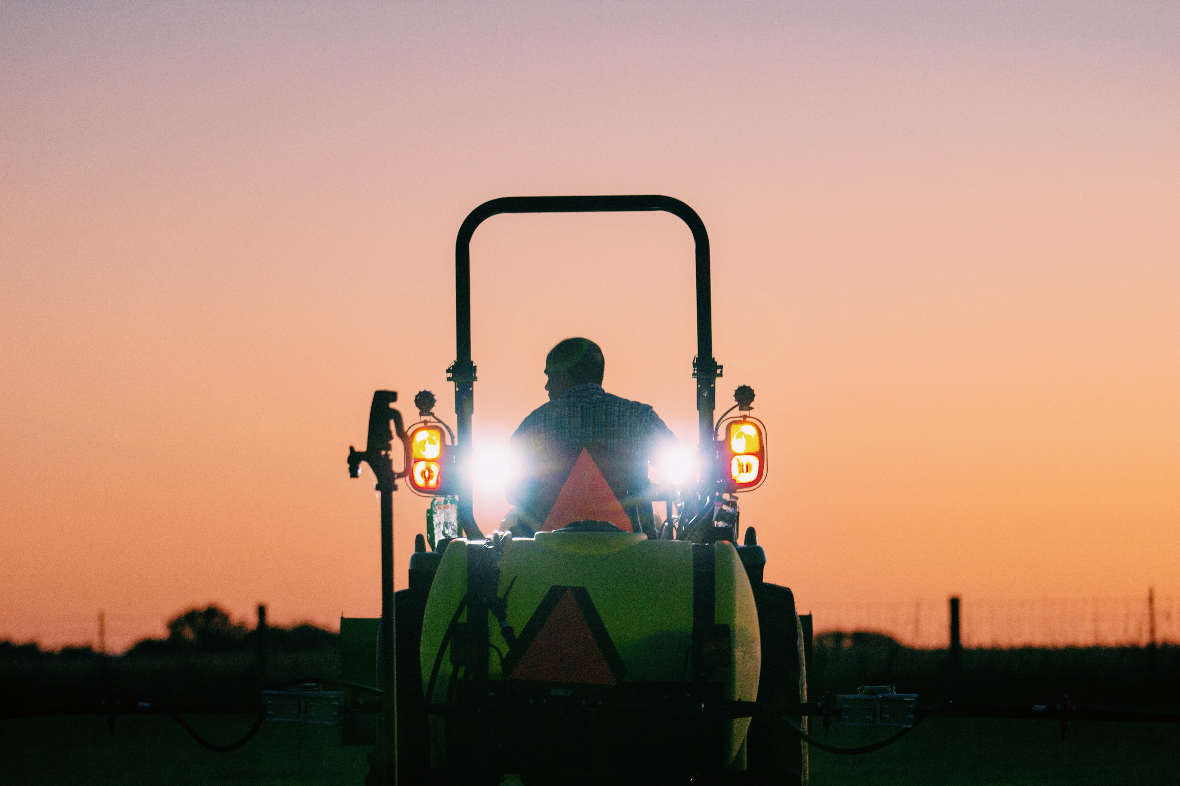 The back of a man on a tractor with a pasture sprayer at sunset
