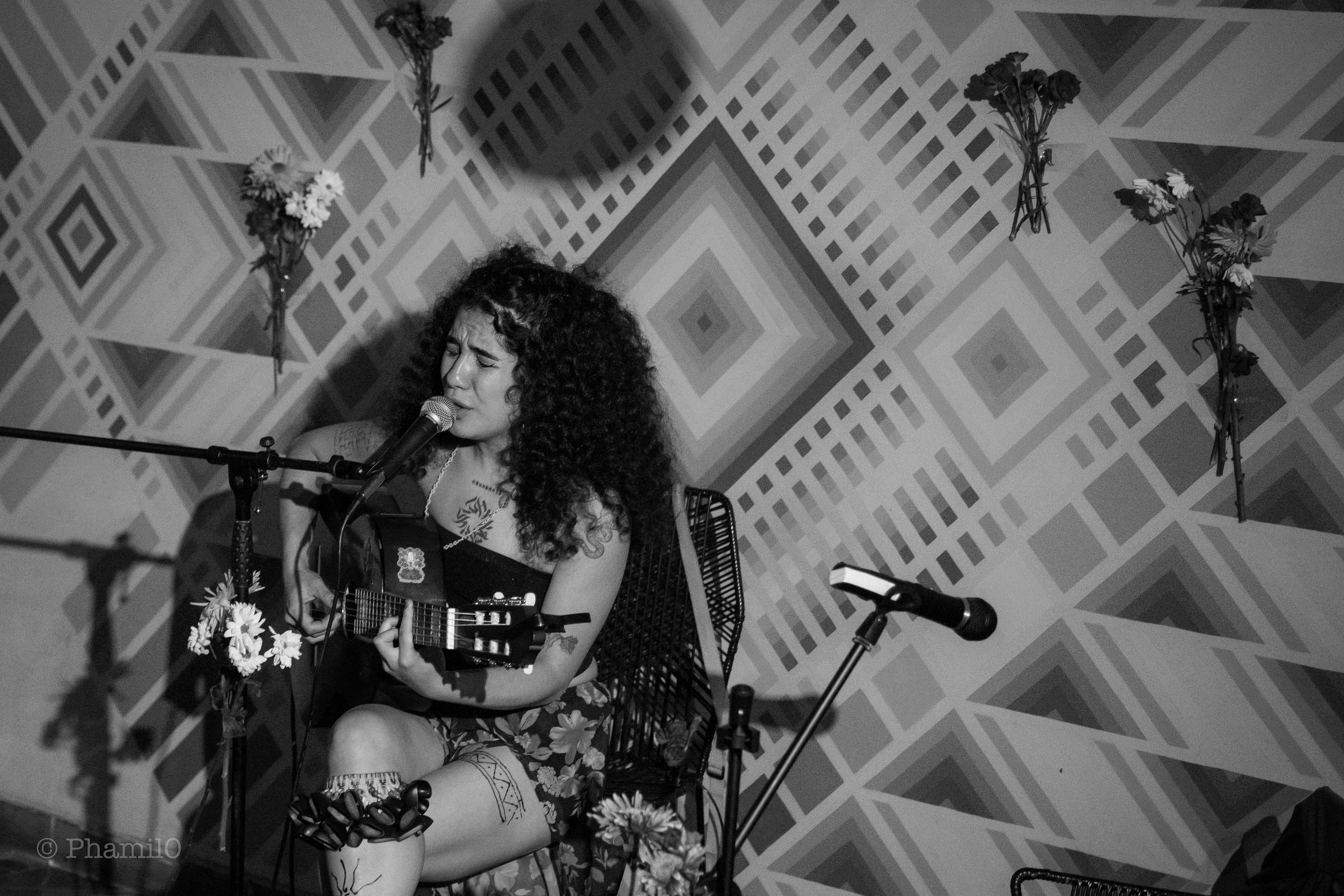 Ana Solarte en un concierto privado en el barrio san Felipe de Bogotá, en el fondo hay rombos, tambien se pueden ber flores pegadas en la pared, y su expresión cuenta una historia  