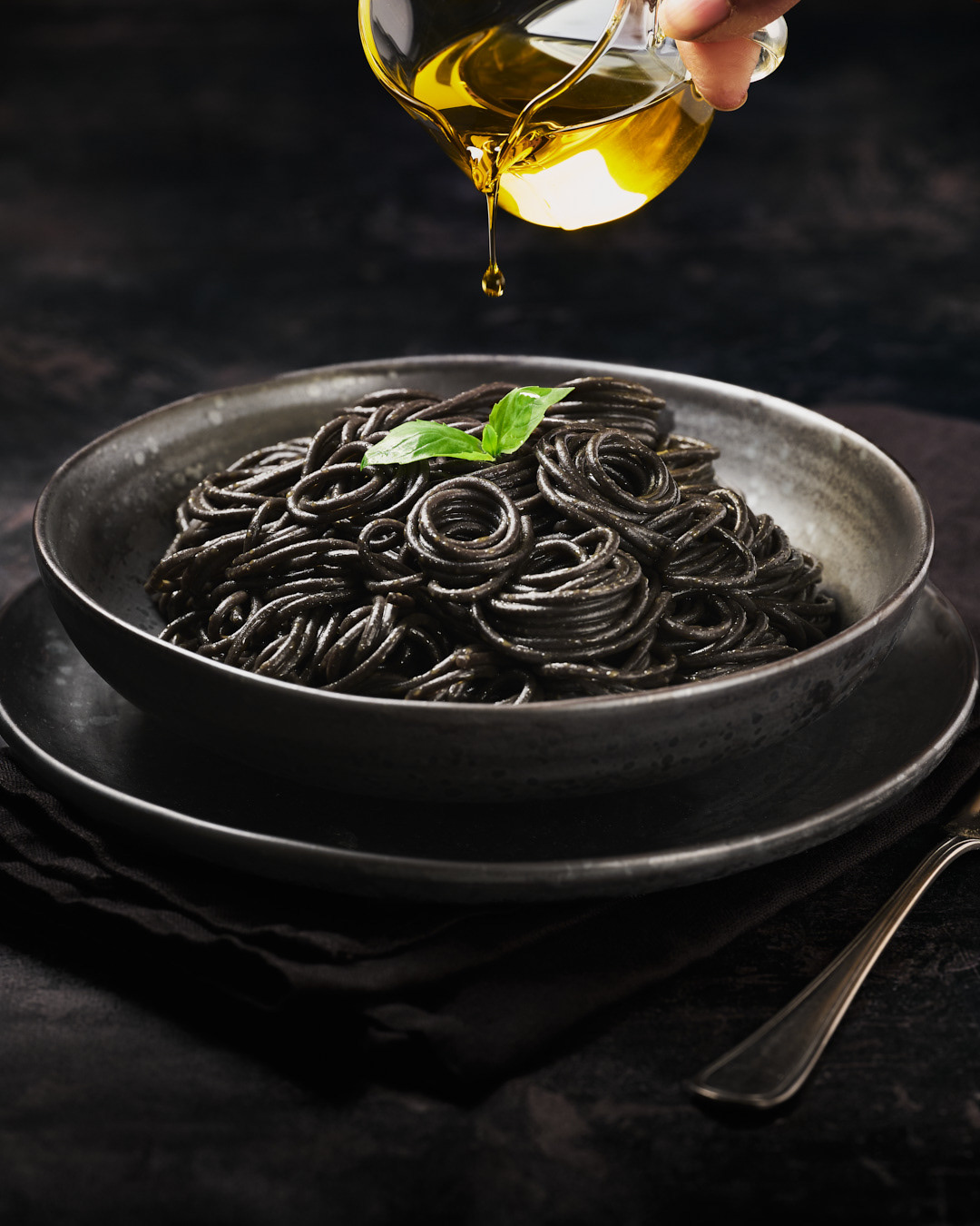 Plate of black spaghetti with a basil leaf and a small jug of olive oil being drizzled over the top