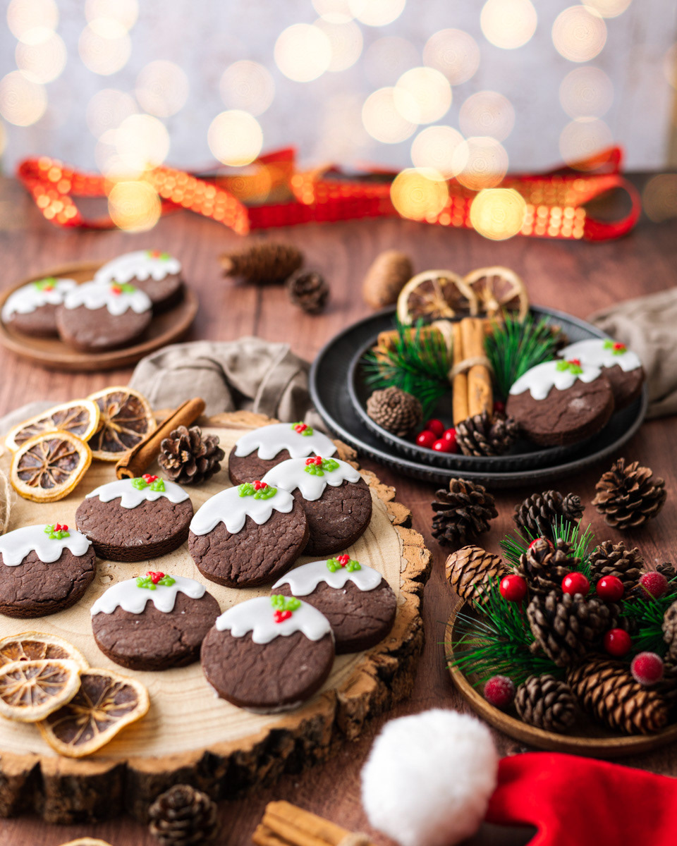 Some Christmas pudding cookies on a wood serving plate with cinnamon and orange slices and Christmas fairy lights by Anusha Stewart