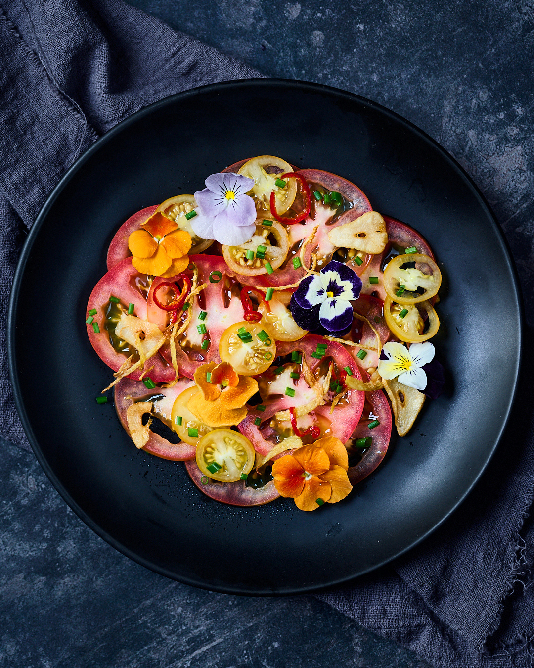 A dark and moody image of a black plate with thinly sliced tomatoes and edible colourful flowers in a dark and moody scene by Anusha Stewart Media