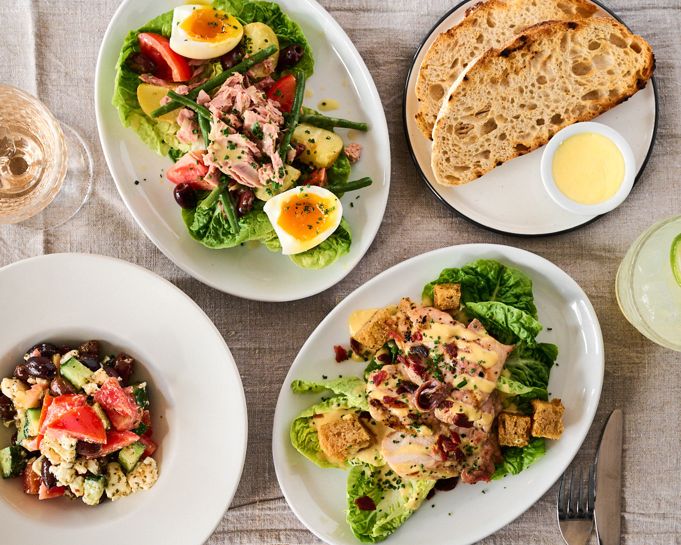 An overhead image of 4 plates of food including a caesar salad, tuna nicoise salad, sourdough and a greek salad by Anusha Stewart Media
