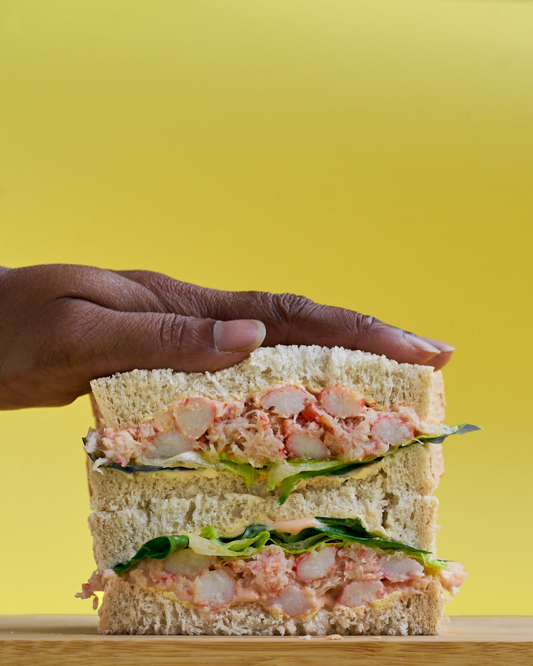 Crab sandwich on a wooden board with a yello background and a hand squashing the sandwich down