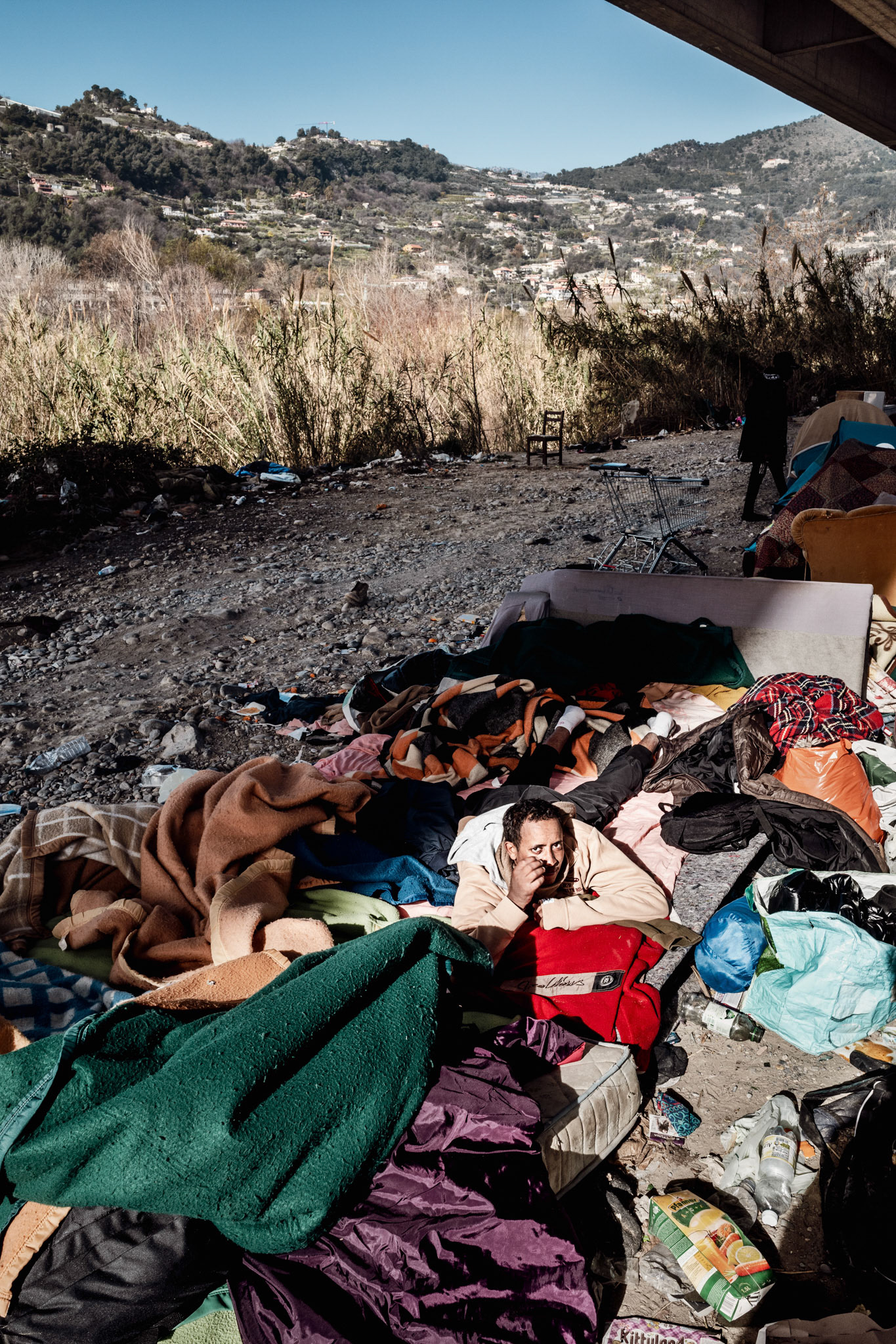 A sunny awakening in the camp under the motorway overpass of Gianchette district, along Roja riverbed. The boy pictured, after a few hours would have crossed the border. The area has been cleared several times in recent years and in November 2020 was completely flooded by storm Alex, as was much of the city centre.