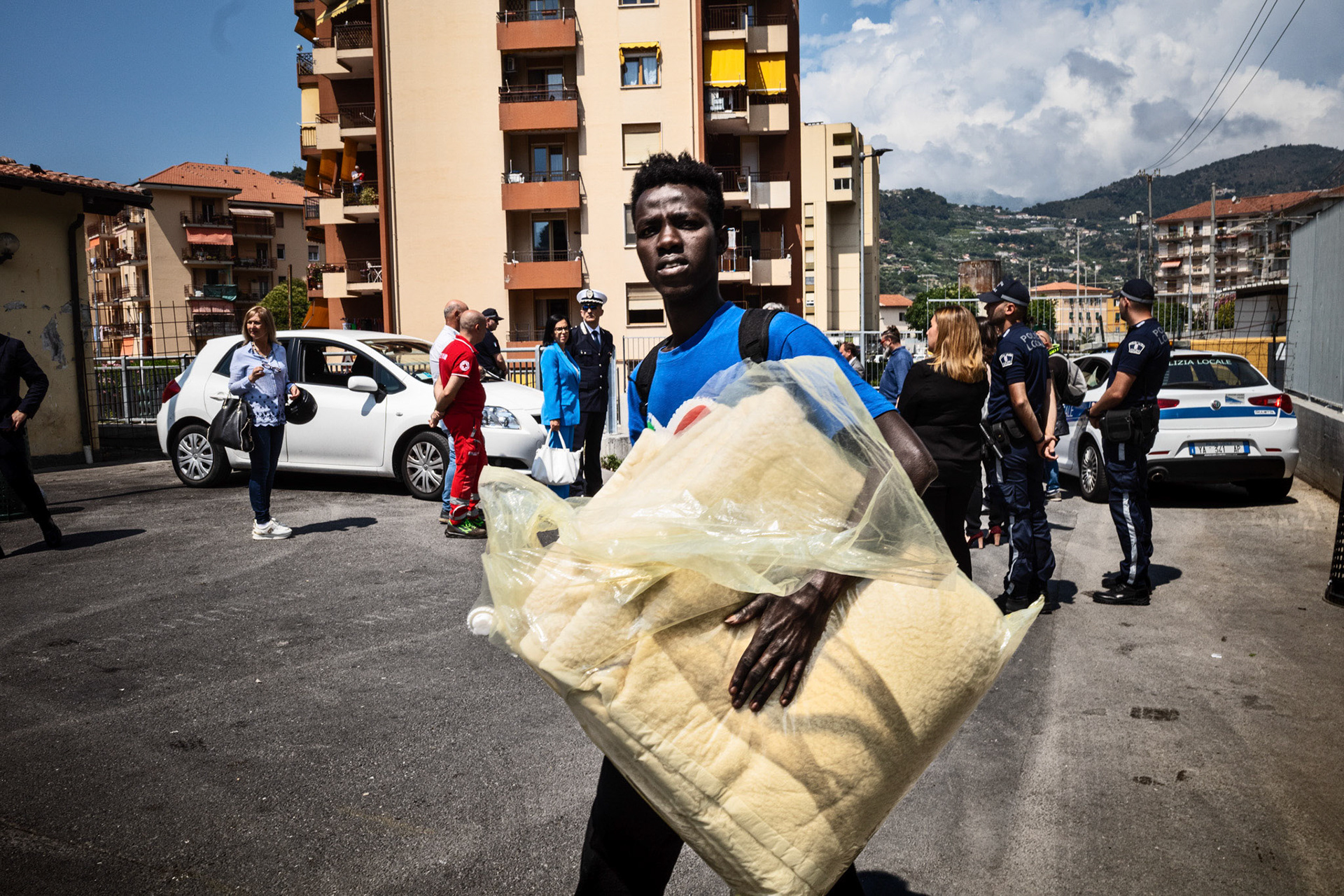 A young migrant has just collected a blanket at the clothing distribution point. Meanwhile, law enforcement officers, journalists, and representatives of the Institutions are awaiting the arrival of the new Prefect