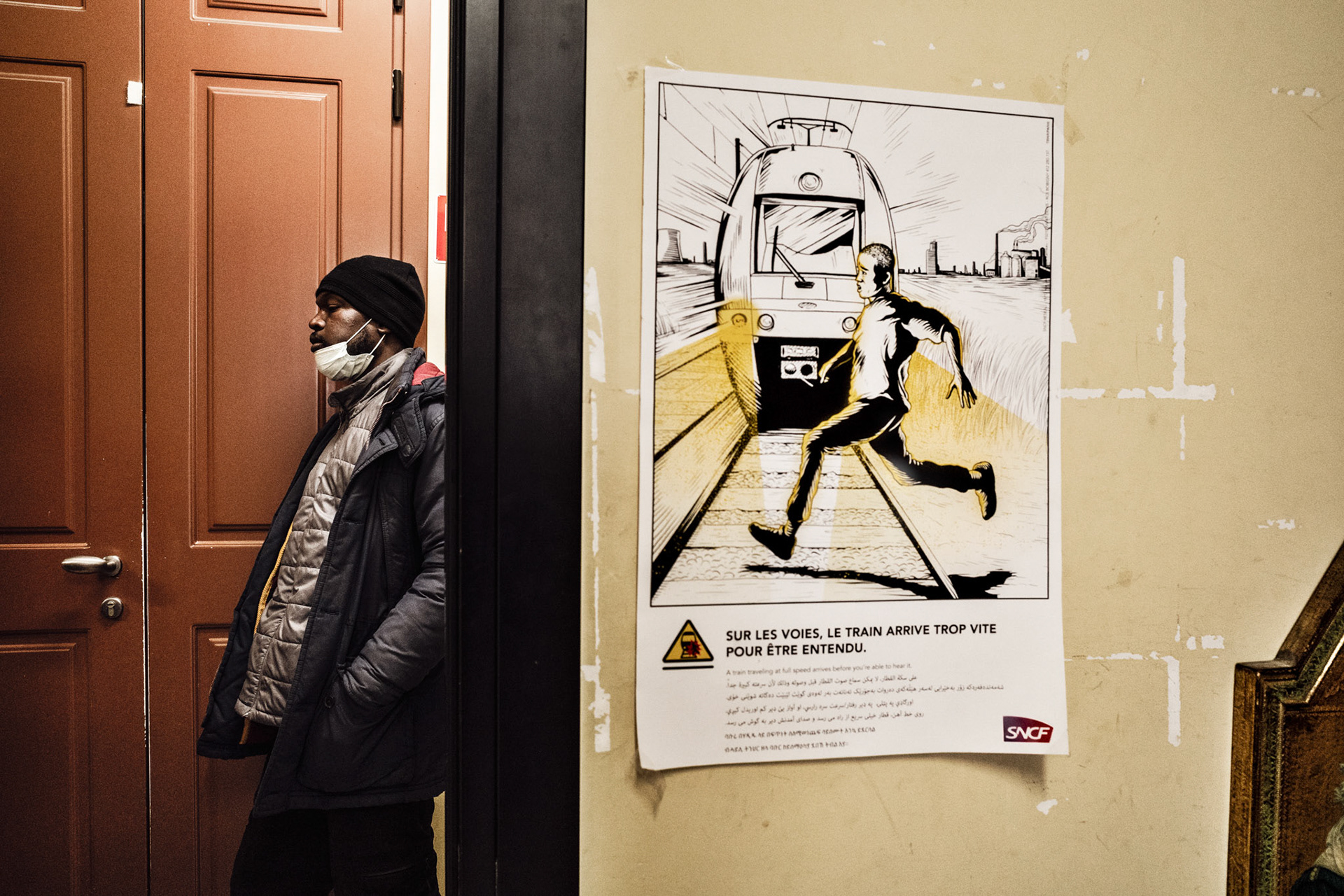 Issa waits his turn at the clothing distribution at Caritas Intemelia. On the wall a poster raising awareness on the risks of crossing the border by SNCF, the French railway company. Since 2015, numerous migrants have died (around 50) or been seriously injured while marching on the railway line or climbing onto the roofs of trains.