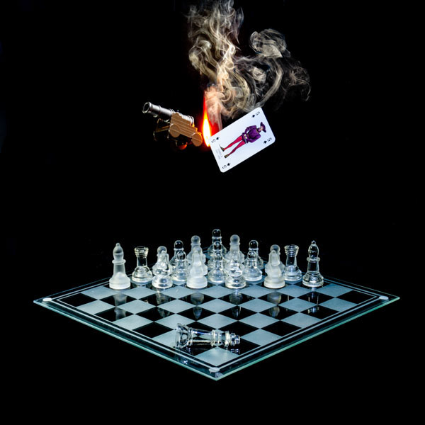 Photographic art print with image of a playing card featuring King James II next to a backfiring cannon represented using a flame and smoke, floating above a glass chessboard with chess pieces strategically place.
