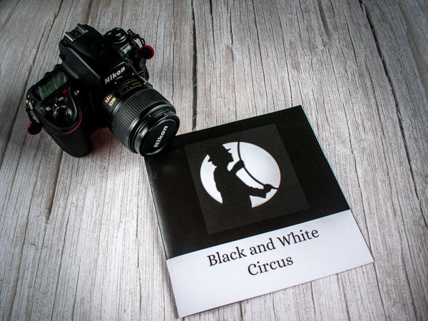 Black and white image of a camera next to a zine called Black and White Circus with a shadow of a figure holding a hoop in a spotlight on the cover.
