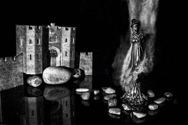 Black and white fine art photograph with a figure representing a witch surrounded by smoke over a fire in front of a castle.
