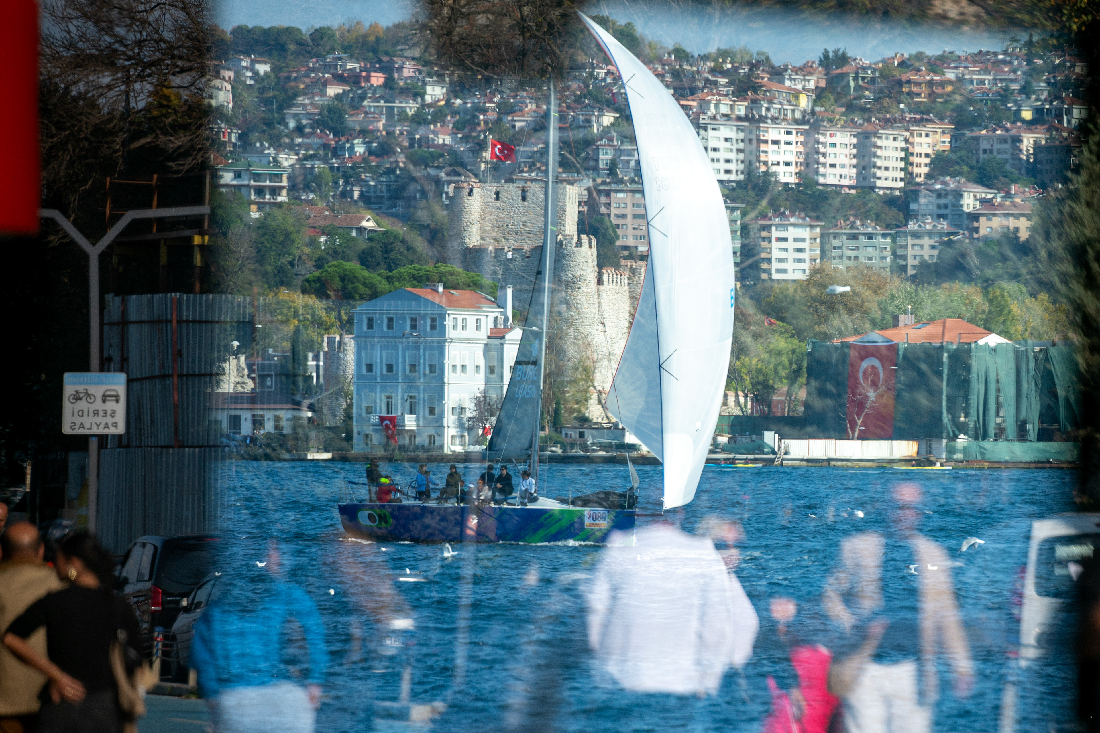 A view of a sailing team competing in the Bosphorus Cup sailing races, with Anadolu Hisarı in the background, as people walk along the Bosphorus on October 29, 2024.