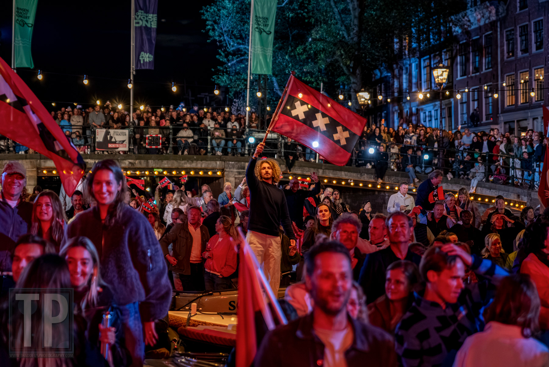 Een man zwaait met de Amsterdamse vlag vanaf een boot, terwijl het publiek toekijkt vanaf de brug en de kade. Prinsengracht, Amsterdam, 22 augustus 2026. · A man waves the Amsterdam flag from a boat as the crowd watches from the bridge and quay. Prinsengracht, Amsterdam, 22 August 2026.