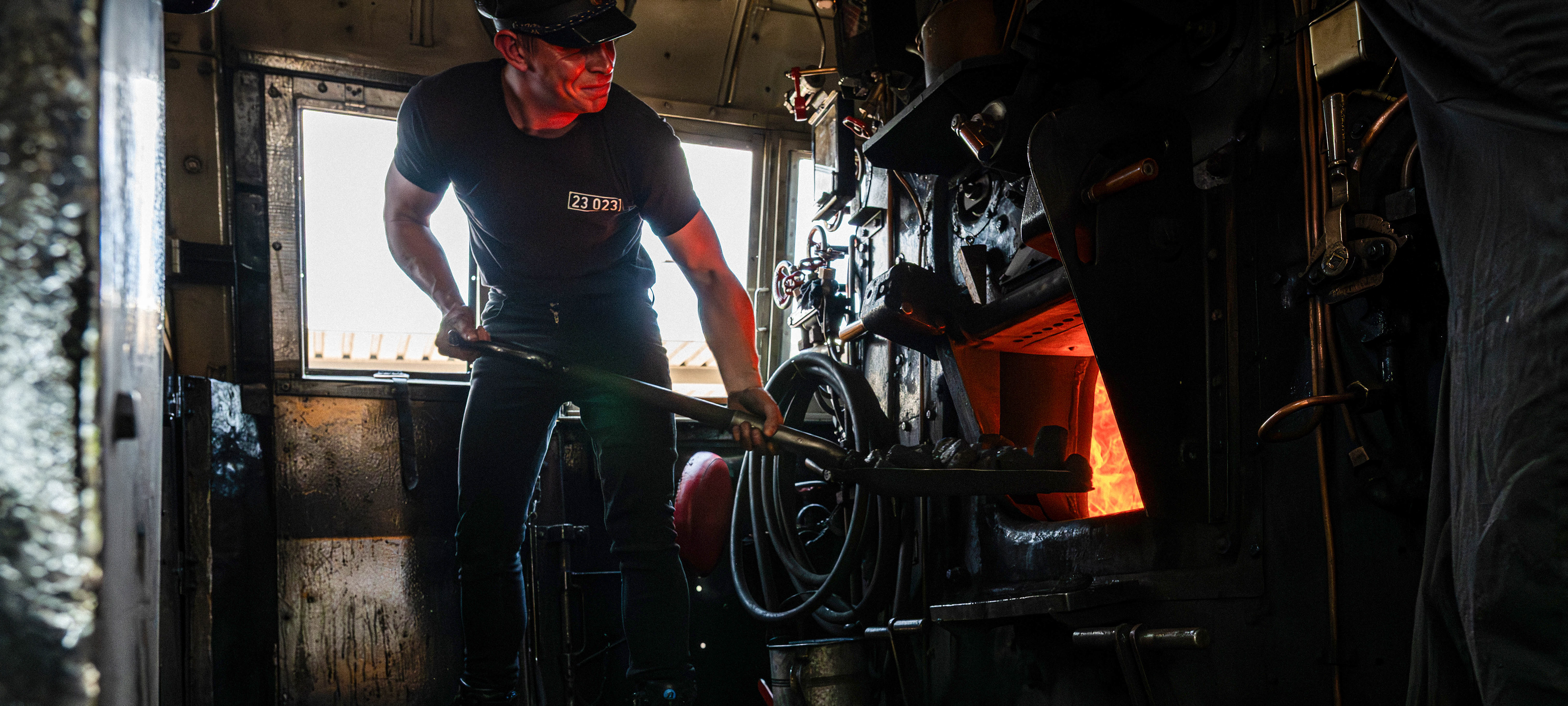 Stoker shovelling coal into the glowing firebox of a steam locomotive, lit red by the fire in the dark engine cab,.