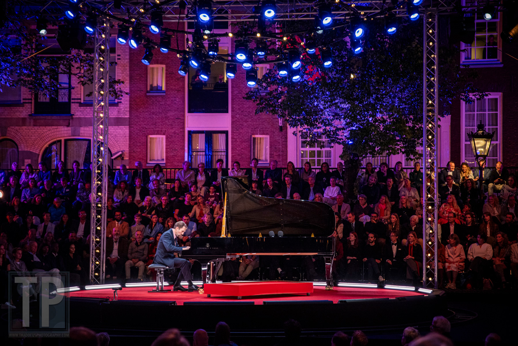 Het ronde podium baadt in blauw licht terwijl pianist Leif Ove Andsnes alleen speelt onder de grachtenpanden in de avondschemering. Prinsengracht, Amsterdam, 22 augustus 2026. · The round stage bathes in blue light as pianist Leif Ove Andsnes plays alone beneath the canal houses at dusk. Prinsengracht, Amsterdam, 22 August 2026.