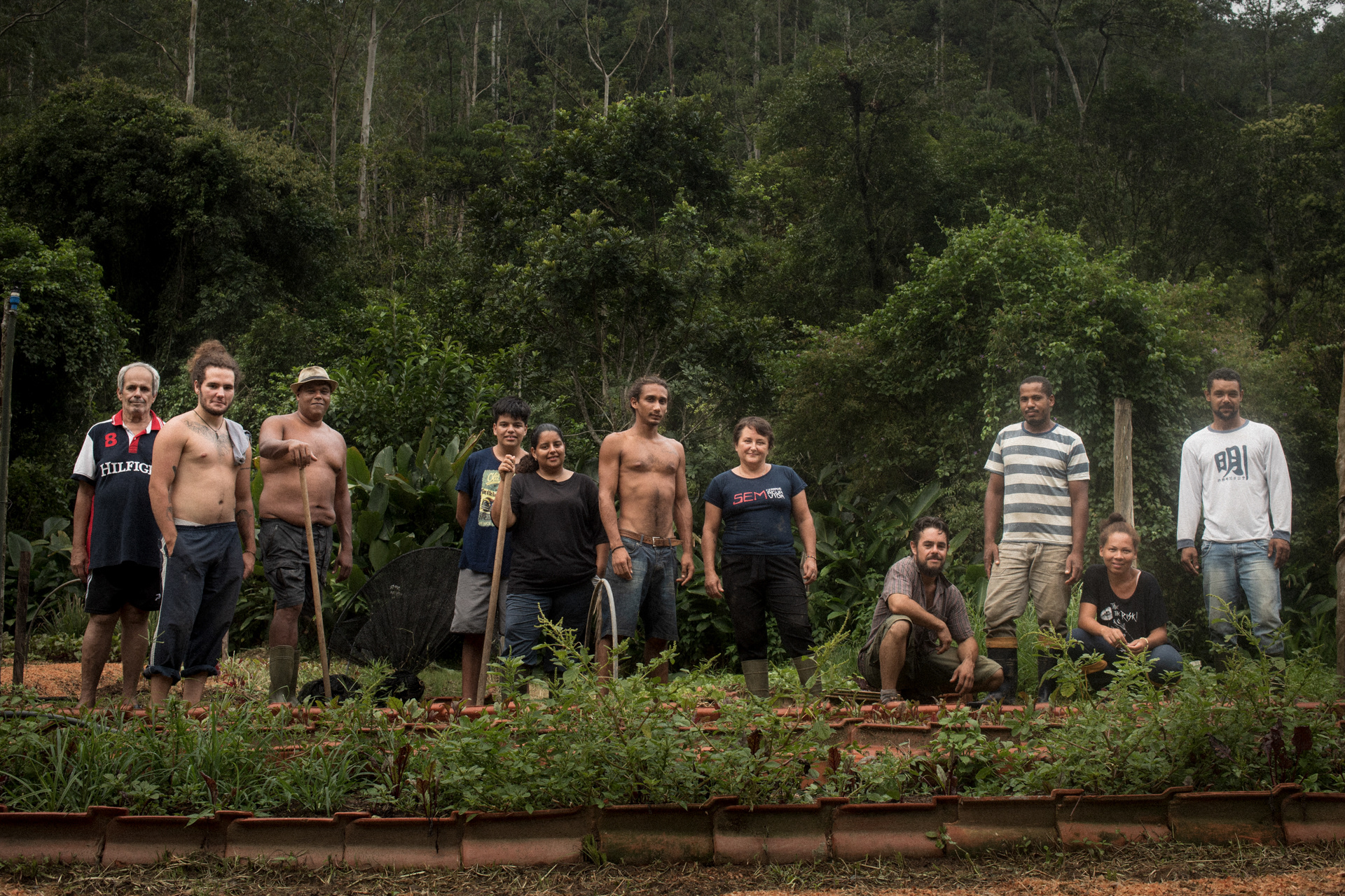 imagem horizontal com onze pessoas lado a lado, algumas em pé, outras agachadas, algumas segurando ferramentas, ao fundo há uma floresta, e estão em uma das horta do estão em frente a uma horta Assentamento Amarildo