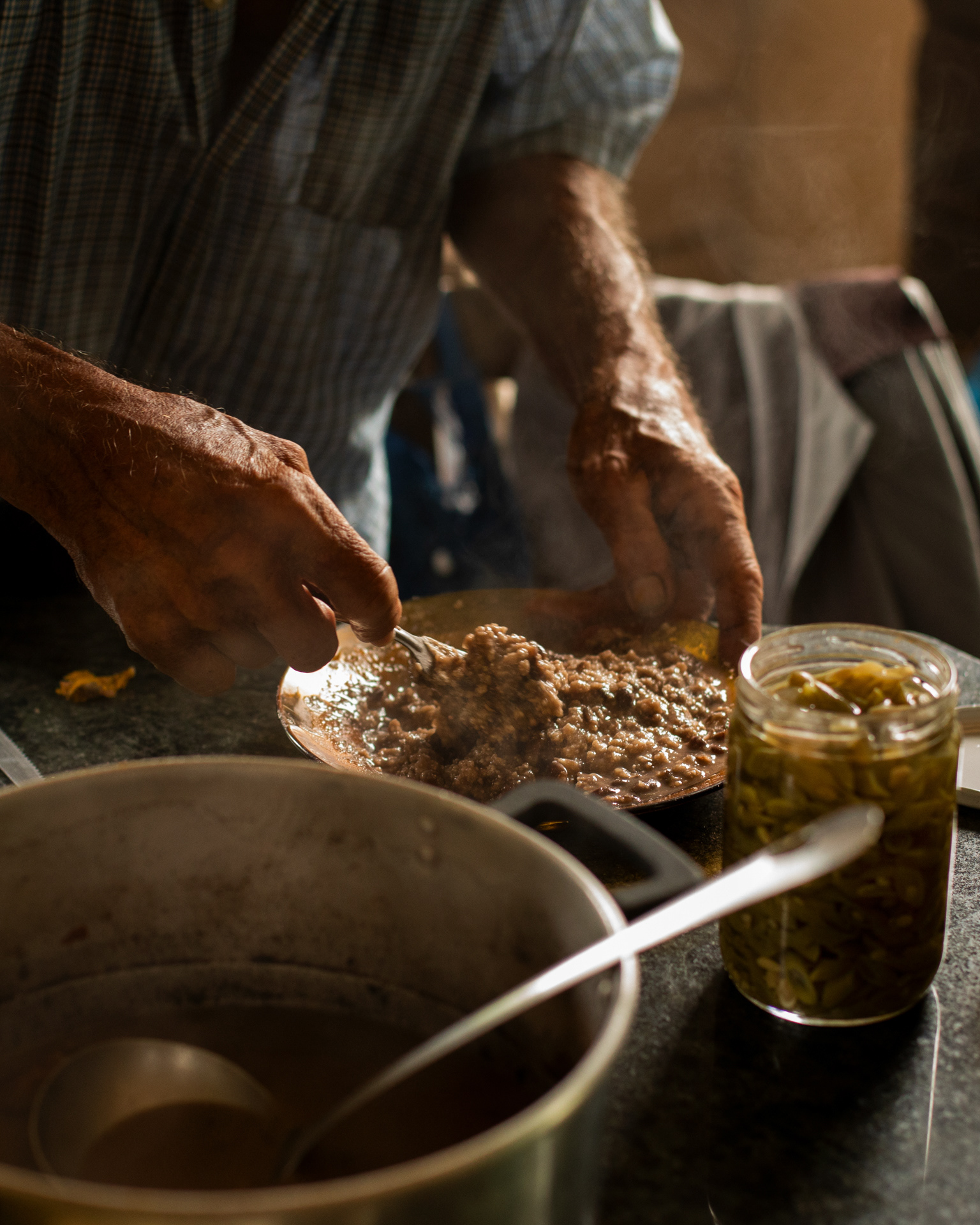 detalhe das mãos de Celso misturando a farinha de mandioca ao feijão para fazer pirão, em primeiro plano vemos a panela e um pote de conserva