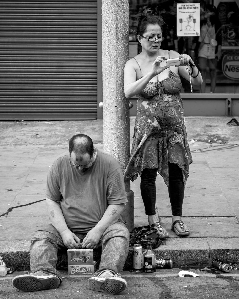 Woman looking at her phone standing beside man eating chicken on the pavement. Image © Nik van Herpt