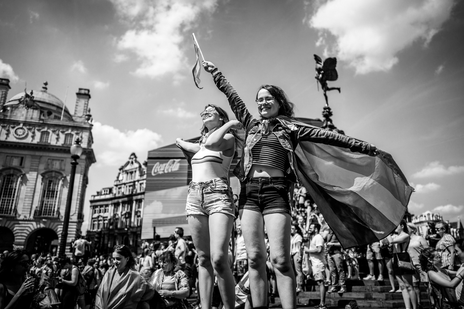 Two girls standing above the crowds waving a Pride flag at Piccadilly Circus. Image © Nik van Herpt
