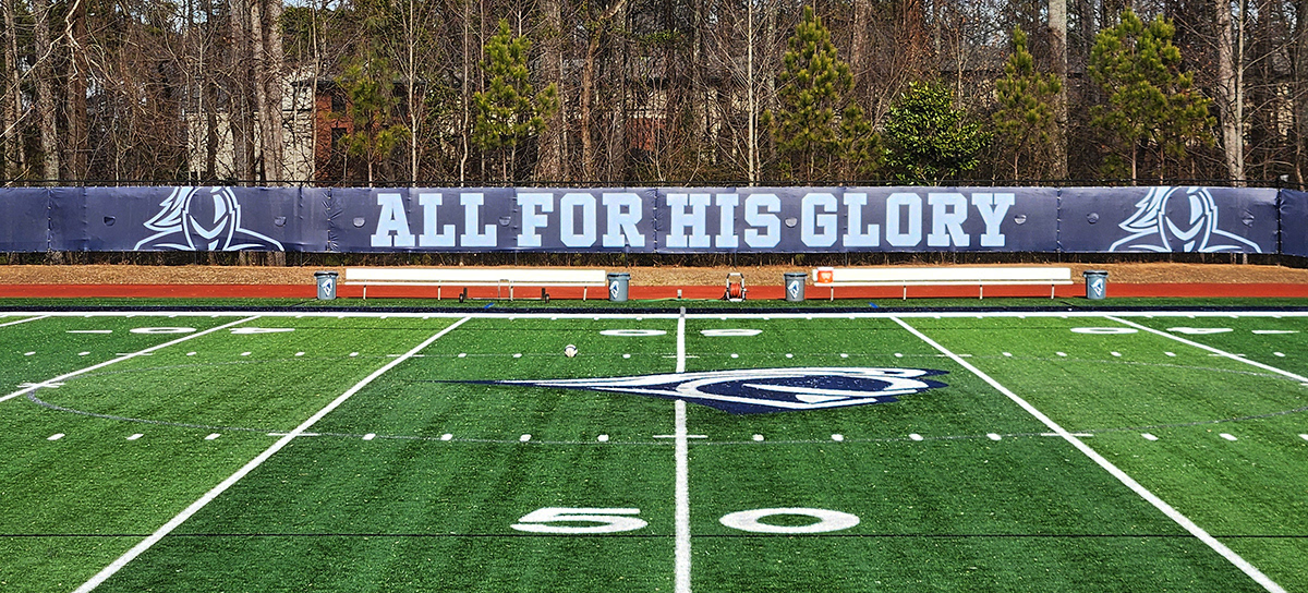 Navy blue fence windscreen panels flank a football field, featuring, from left to right in white, an outline of a knight, the words "ALL FOR HIS GLORY," and another knight outline.