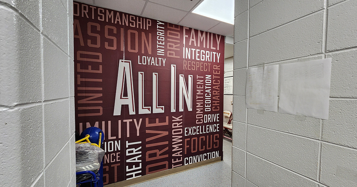 An adhesive wall mural with a burgundy background, featuring white and lighter shades of burgundy lettering in the foreground. The word cloud includes the words: sportsmanship, passion, loyalty, pride, integrity, family, respect, commitment, dedication, character, teamwork, focus, conviction, heart, united, and "all in," with "all in" being the largest font and positioned in the center of the word cloud.