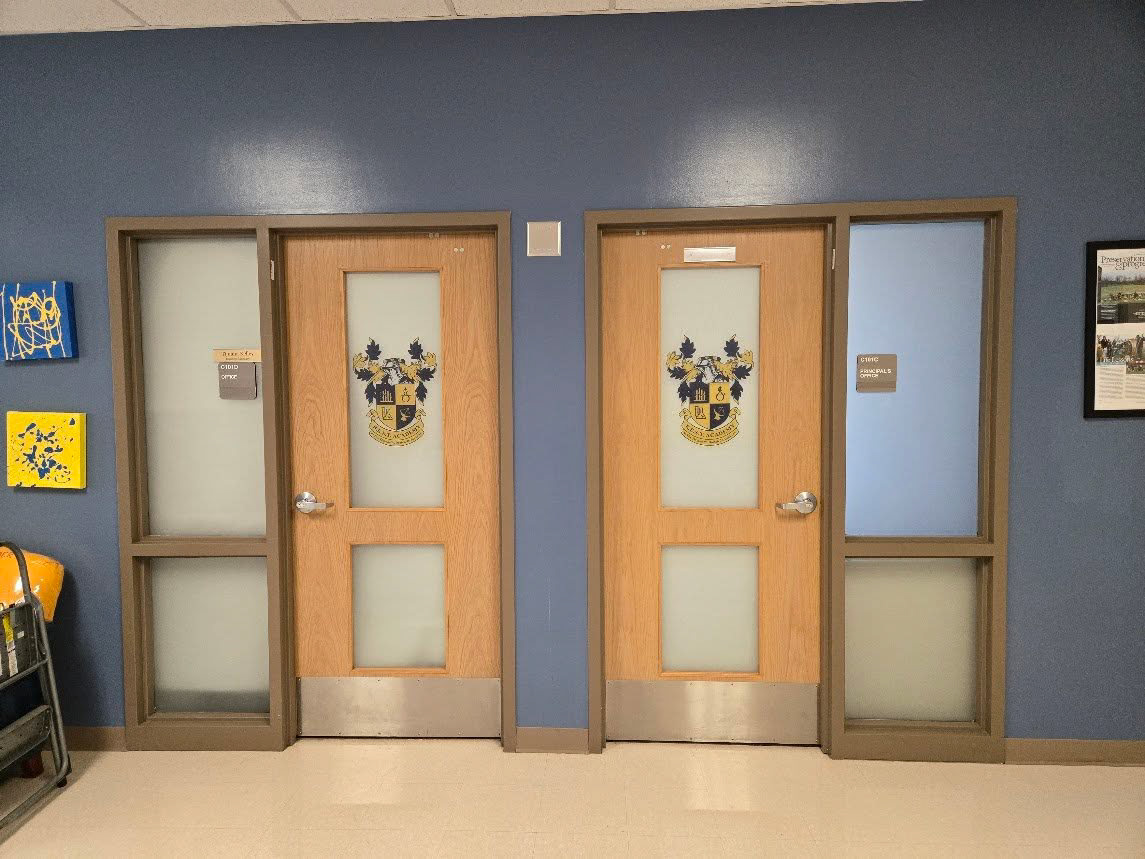 Straight-on view of two interior school doors with window panels, flanked by large split windows on each side. All windows feature frosted, perforated film, while the top windows of the center doors display a custom yellow-and-navy-blue school crest.