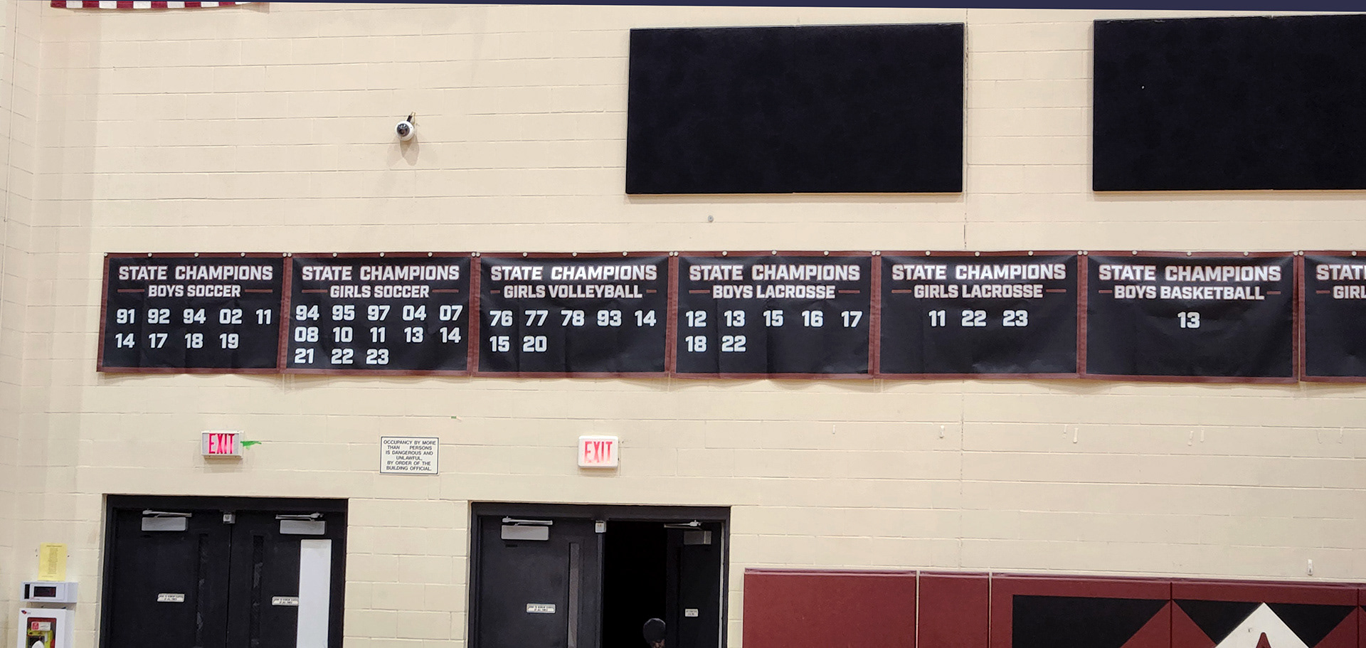 A black 10oz VCP windscren rectangular banner with a maroon border, white text and numbers promoting state champions for Wando High School. The banner is displayed on the upper part of the gym walls, just above the door