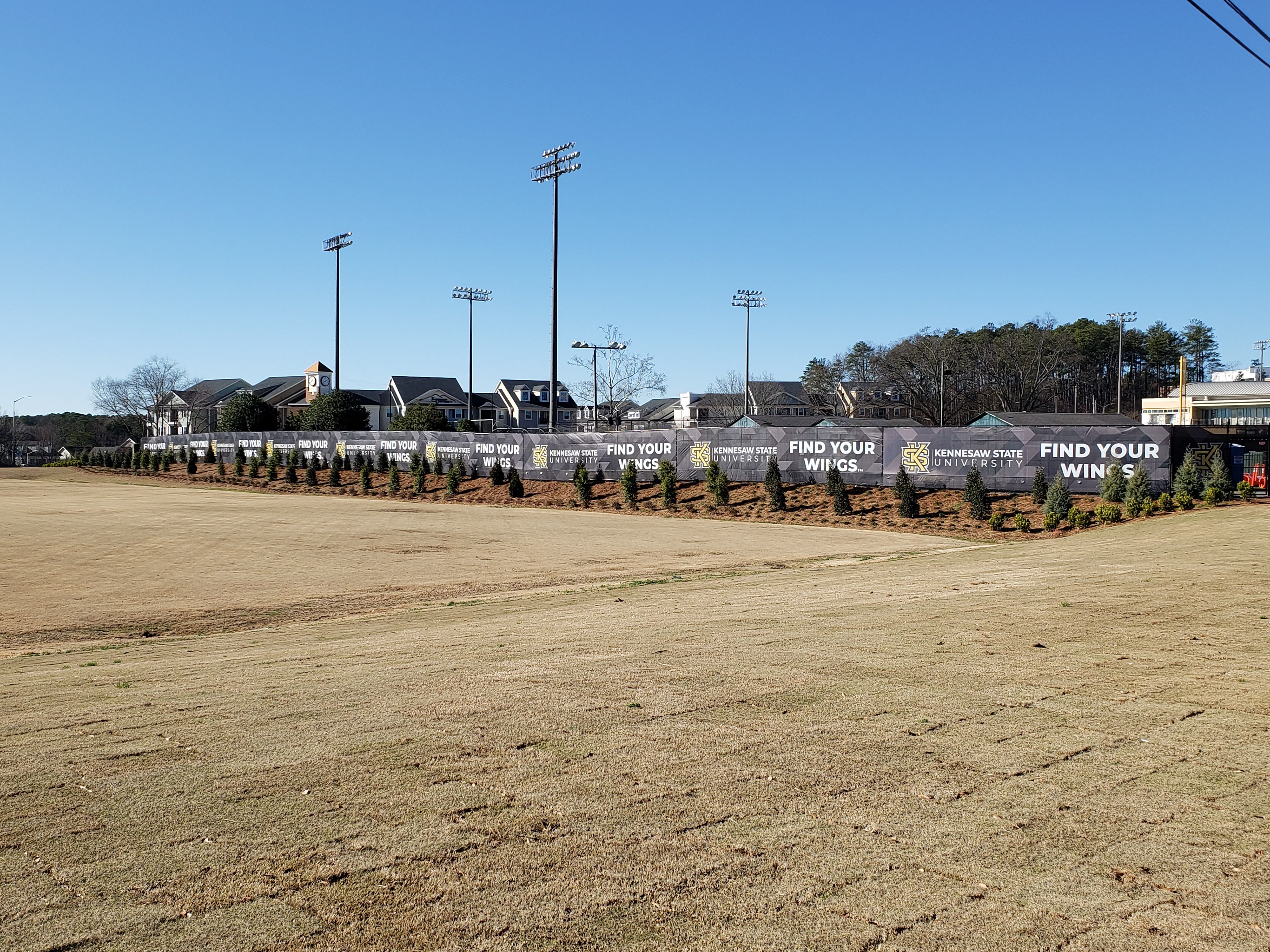 A series of printed windscreens attached to a fence around a field, featuring a geographic print under the printed words and logos. The first panel displays the words 'FIND YOUR WINGS' in white. The second panel shows 'KENNESAW STATE UNIVERSITY' in white, alongside a 'KS' logo in yellow outlined in black. This print pattern repeats multiple times along the fence.