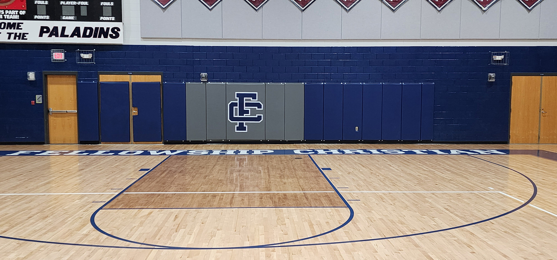 Sixteen vinyl-covered backstop pads in a basketball court gym, attached to a wall. The four pads on the left and the seven pads on the right are blue, with two of the left pads placed on wooden doors. The five middle pads are gray, each featuring a blue 'FC' monogram logo outlined in white printed in the center.