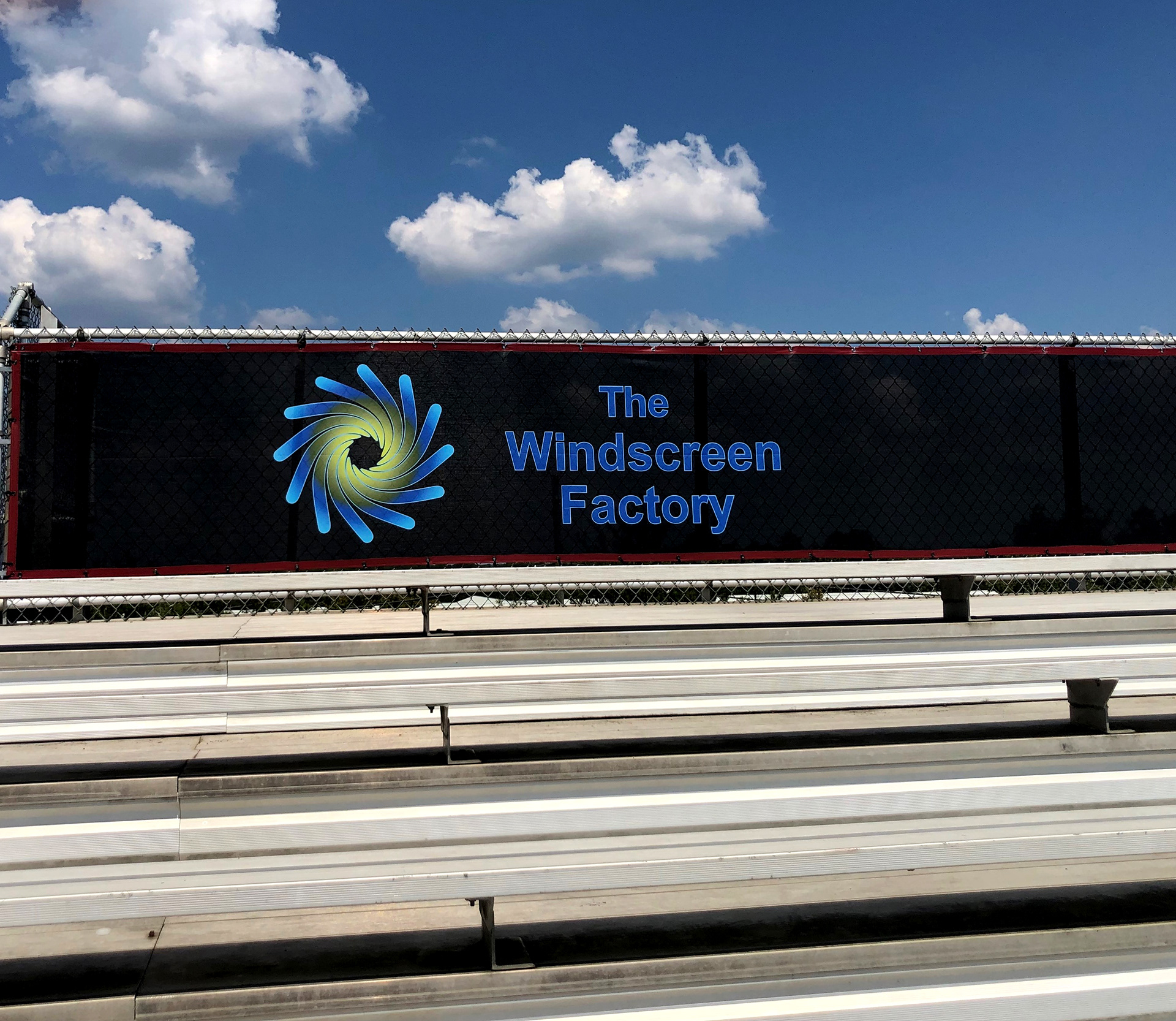 A branded windscreen panel attached to the back railing of bleachers, viewed from a slightly left angle. The windscreen features the text 'The Windscreen Factory' in blue, to the right of the company's logo.