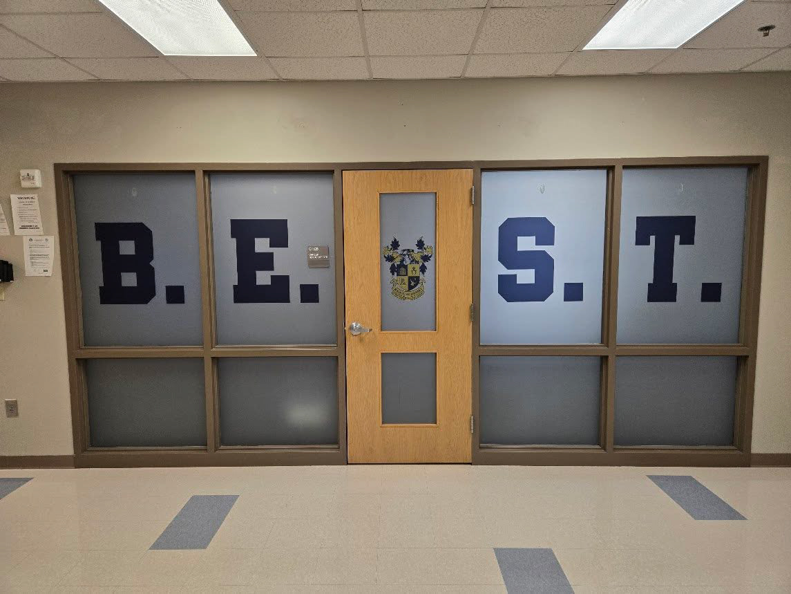 Straight-on view of an interior school doorway with a windowed door and surrounding windows. Navy blue letters spelling ‘B E [school crest] S T’ appear across the top windows from left to right, with a yellow and navy blue school crest centered on the door. Remaining window areas are frosted.