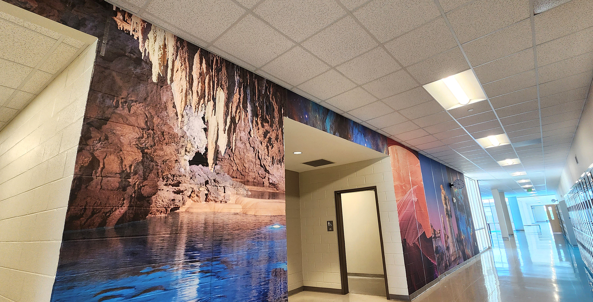 A floor-to-ceiling adhesive vinyl wall mural in a hallway with an intersecting hallway to the left and an alcove with access to two bathrooms near the middle. The mural features, from left to right, an image of a lake inside a cave, an image of colorful sand and rock formations, and beyond that, the images are too distant to discern clearly. 