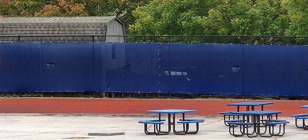 A Navy blue fence windscreen separates picnic tables beside the track and football field from an outside pedestrian area.