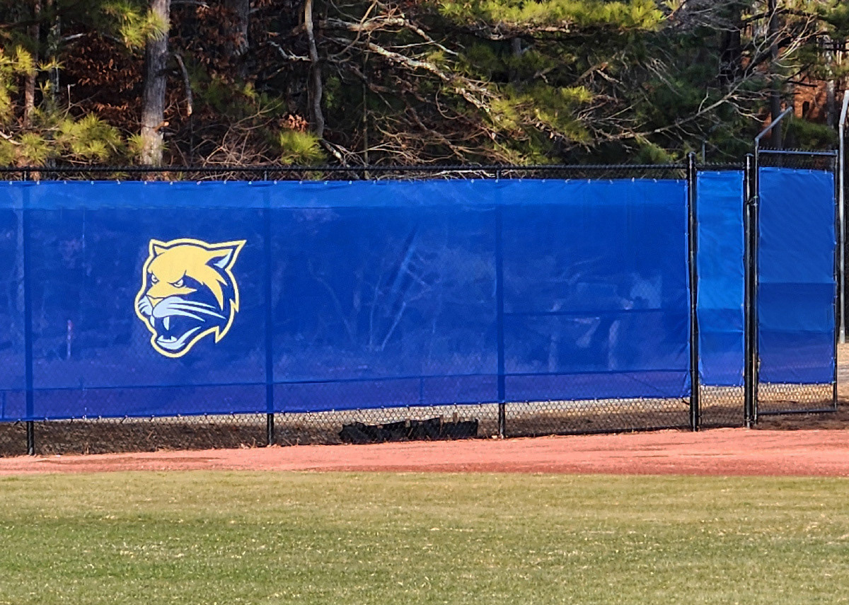 blue windscreen attached printed with a yellow and white cougar logo attached to a fence barrier on the outter edge of a baseball field