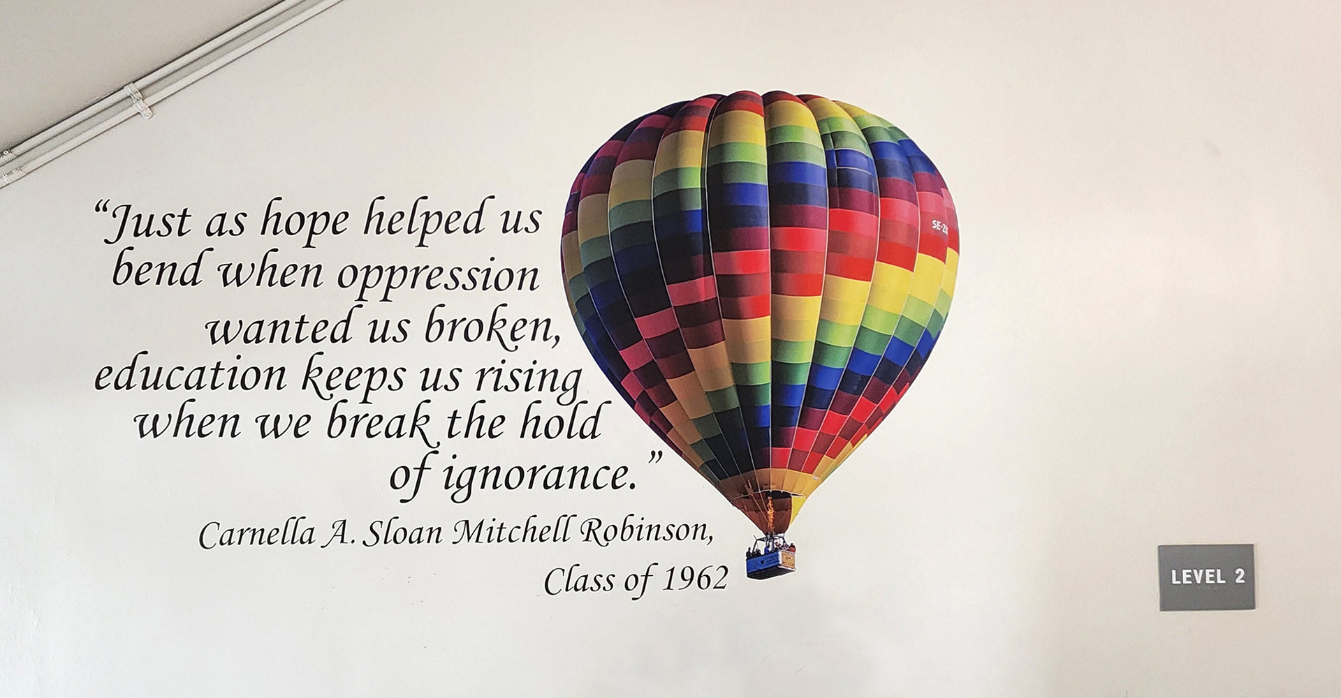 On a wall near a stairwell landing or platform, an adhesive vinyl image of a multi-colored rainbow hot air balloon, alongside the quote 'Just as hope helped us bend when oppression wanted us broken, education keeps us rising when we break the hold of ignorance' – Carnella A. Sloan Mitchell Robinson, Class of 1962. 