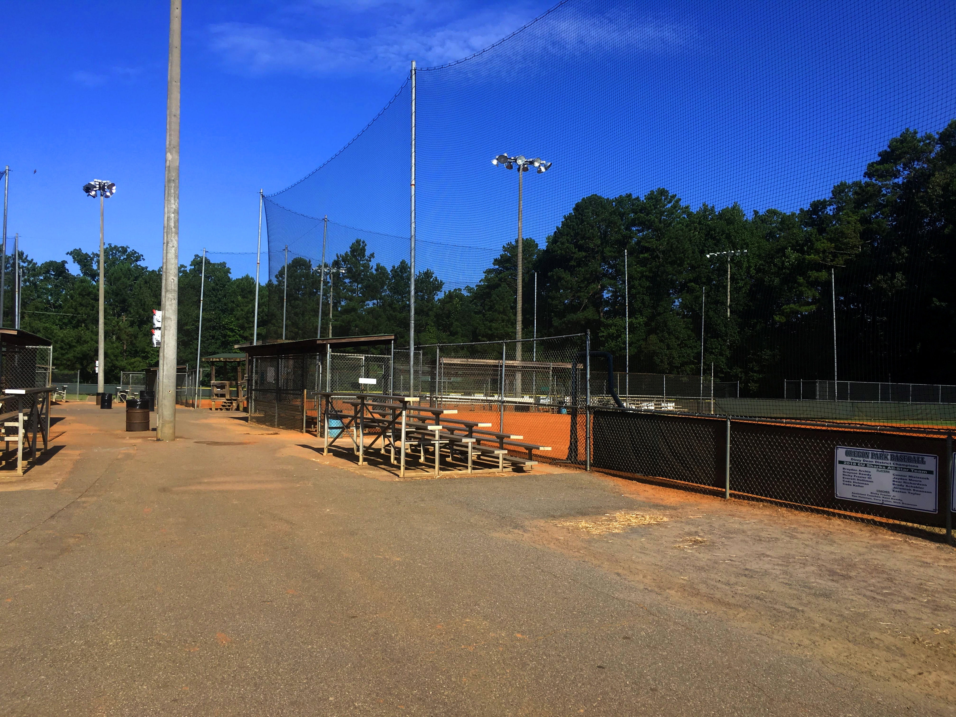 To the right, vertical sideline sports netting surrounds a baseball field. Next to the field, there is a set of metal bleachers adjacent to a baseball dugout. On the left, a walkway for visitors and spectators runs between sports fields, with another set of bleachers partially visible to the far left. In the distance, a third baseball field is visible center front. 