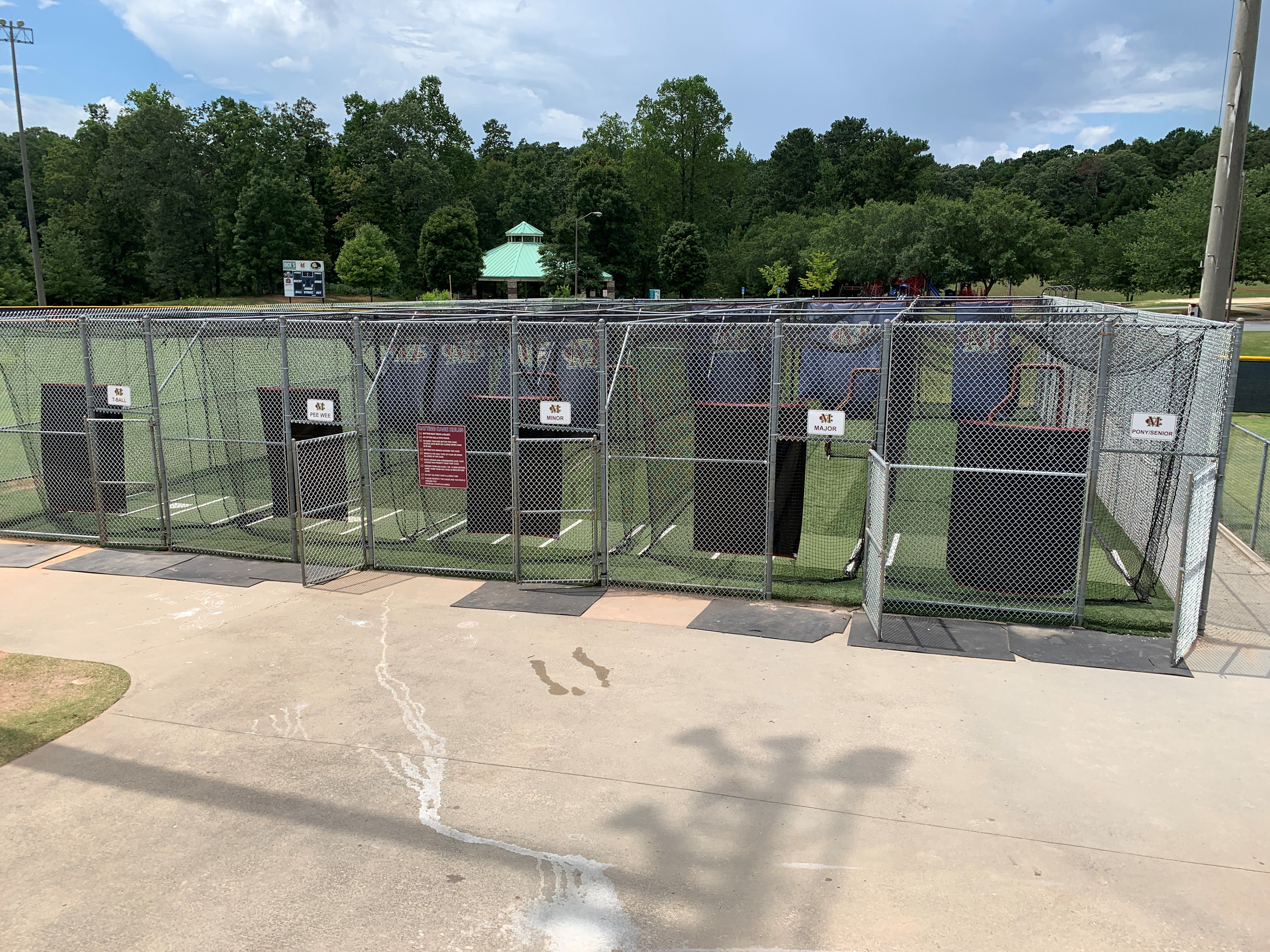 In the foreground a view of a silvertone metal baseball batting cage from the outside, with black sports netting separating the lanes and lining the cage, and green turf covering the ground. In the background, part of a sports field is visible.