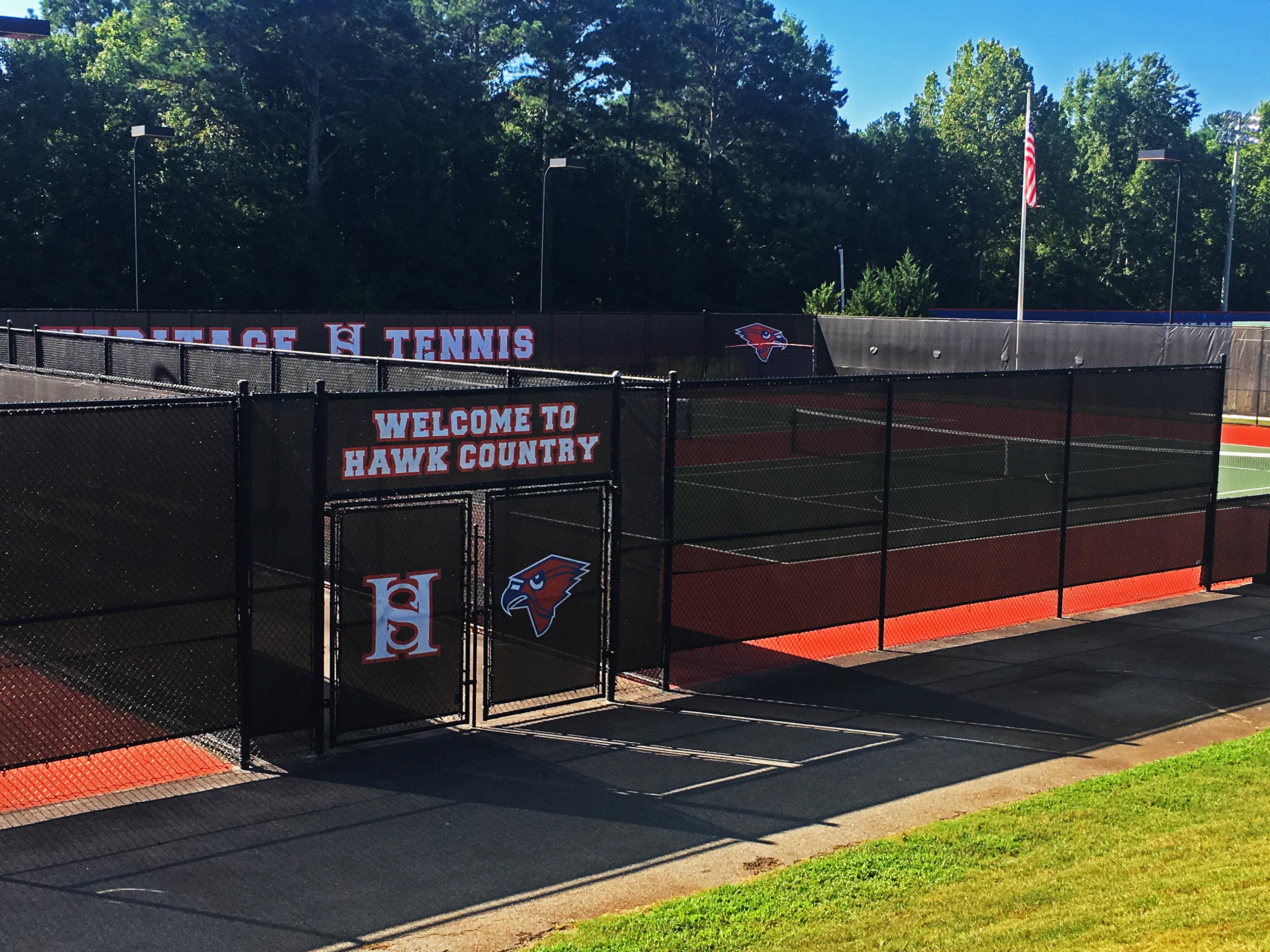 Branded black fence windscreen surrounding separate tennis court areas, including over and on the entry doors. Viewed from inside the tennis courts, to the back right, the windscreen displays 'HERITAGE HS TENNIS,' with the 'HS' monogram logo showing the two letters as one, and a red cardinal head logo with a black beak, outlined in black and again outlined in white. Above the entrance to the tennis court, the windscreen features the words 'WELCOME TO HAWK COUNTY' in white, outlined in red. On the door to the left, there is a 'HS' monogram logo in white outlined in red, while the door to the right displays the red cardinal head logo with a black beak and black outline, then outlined again in white.
