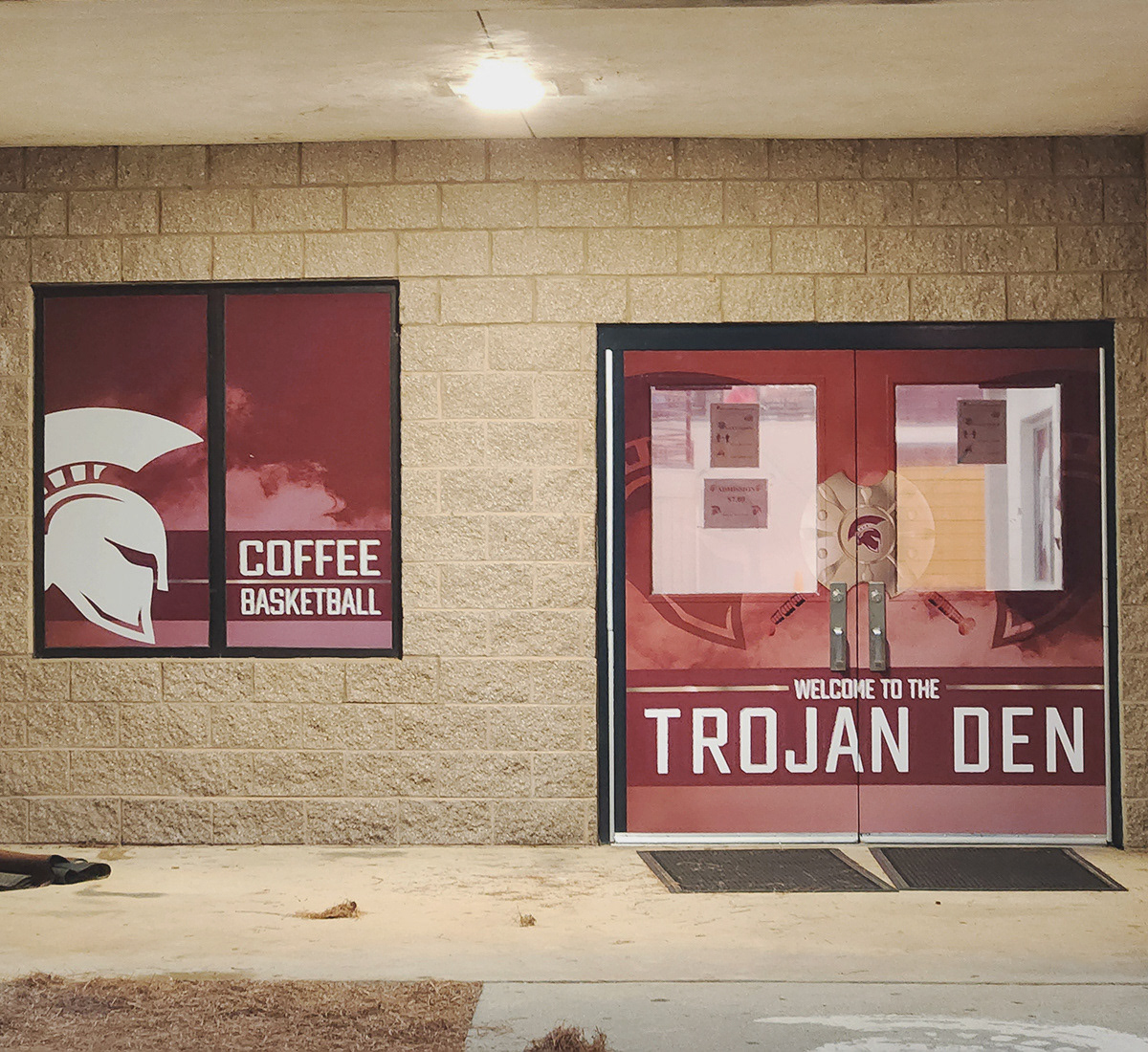 Standing outside the entrance to a school building, with two doors on the right and a double-pane window on the left. The doors are covered with full-print adhesive vinyl, and the windows at the center top of the doors are covered with window perf. Together, the vinyl and window perf create an image of two darker burgundy Trojan helmet silhouettes facing each other, with a Trojan metal shield in the middle. A dark burgundy banner beneath the shield reads 'WELCOME TO THE TROJAN DEN' in white text, with a burgundy smoke background.