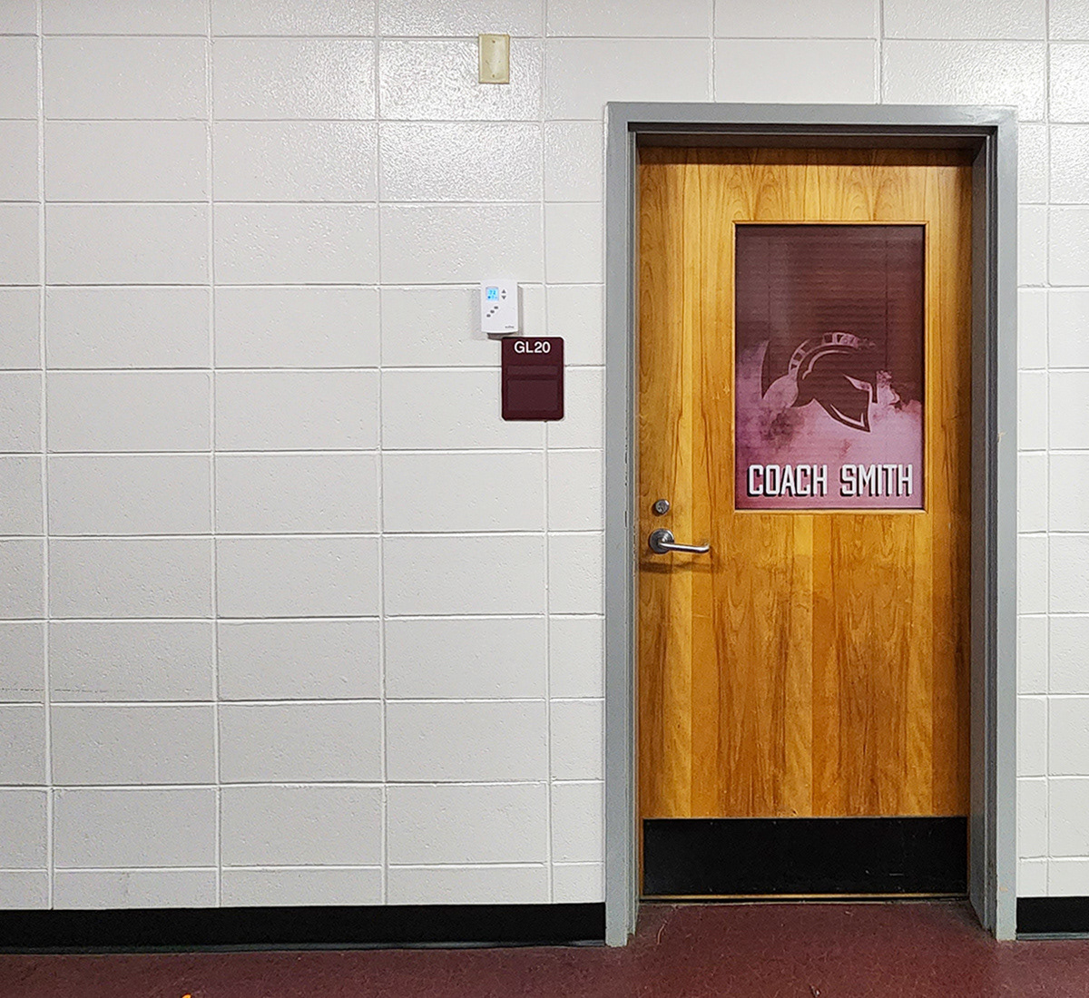 A hallway in a school, with a door to the high school's coach's office. On the top center of the door is window perf print with a burgundy smoke background, a darker burgundy Trojan helmet silhouette, and white text reading 'COACH SMITH