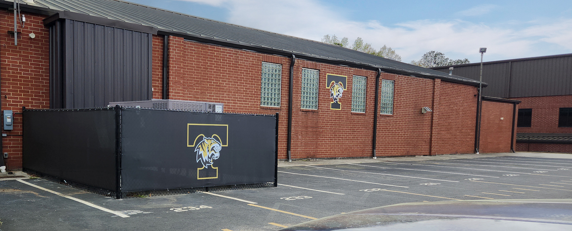 Black windscreen branded with the 'tiger nation' logo attched to fencing surrounding an large AC unit near a parking lot and a large brick building branded with an adhesive vinyl 'tiger nation' logo.