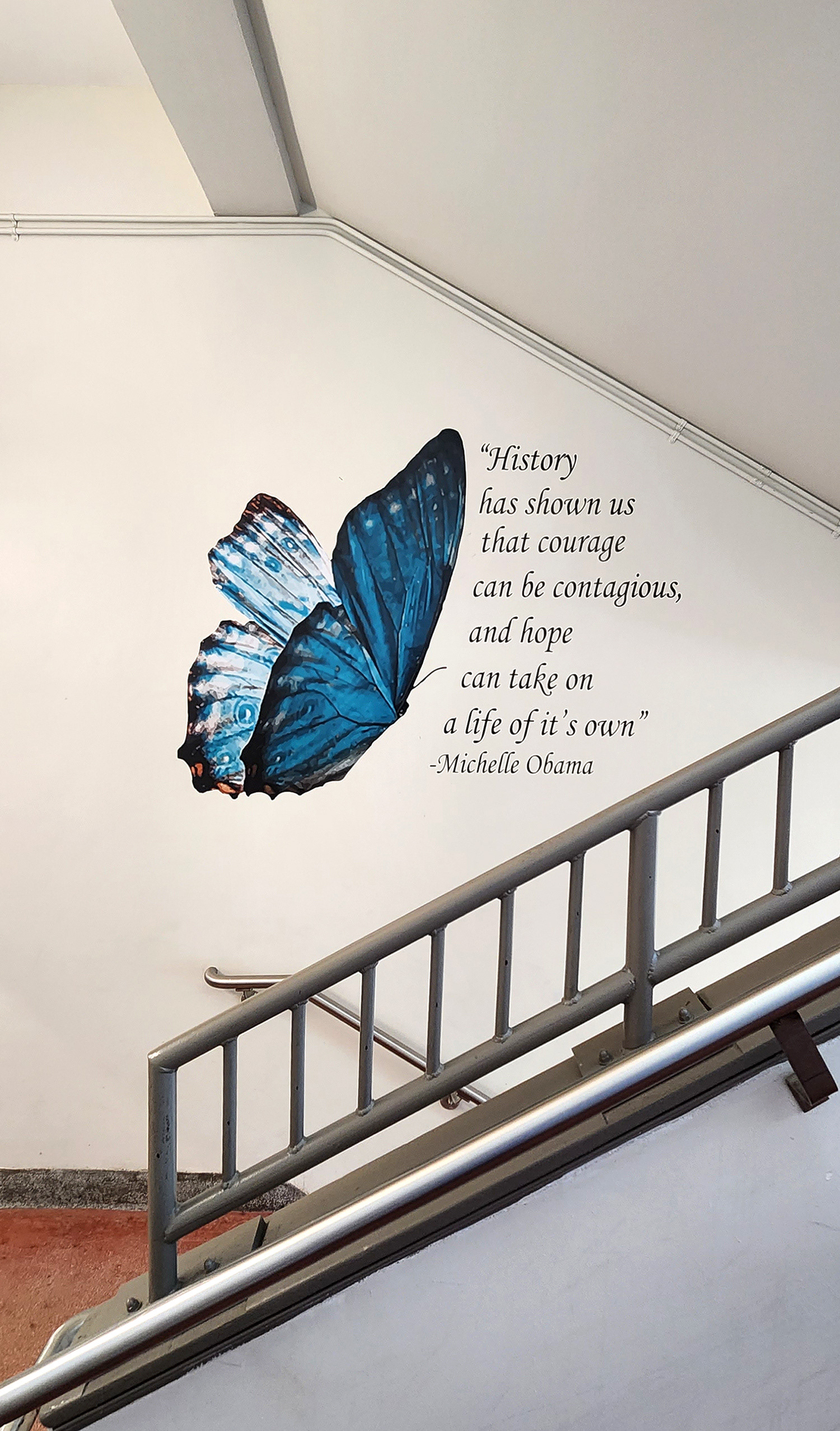 On a wall near a stairwell landing or platform, a blue butterfly adhesive vinyl decal is positioned to the left of cut adhesive vinyl black lettering that reads 'History has shown us that courage can be contagious, and hope can take on a life of its own.' - Michelle Obama